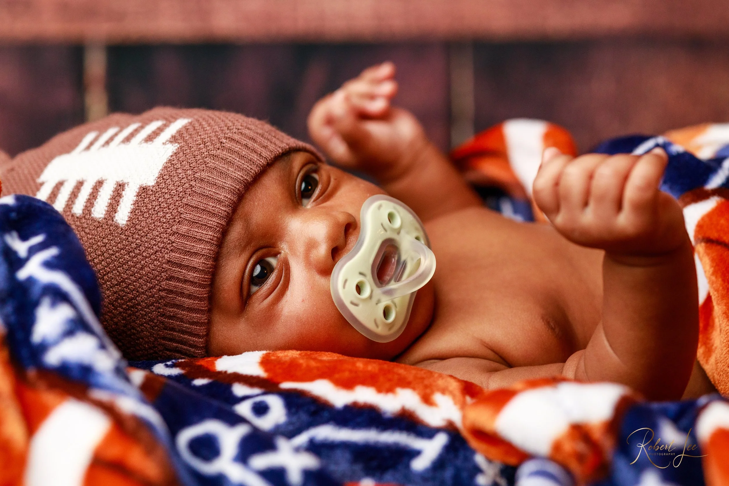 Close-up of a young baby lying down, wearing a knitted brown hat, with a pacifier in their mouth, wrapped in a colorful blanket.