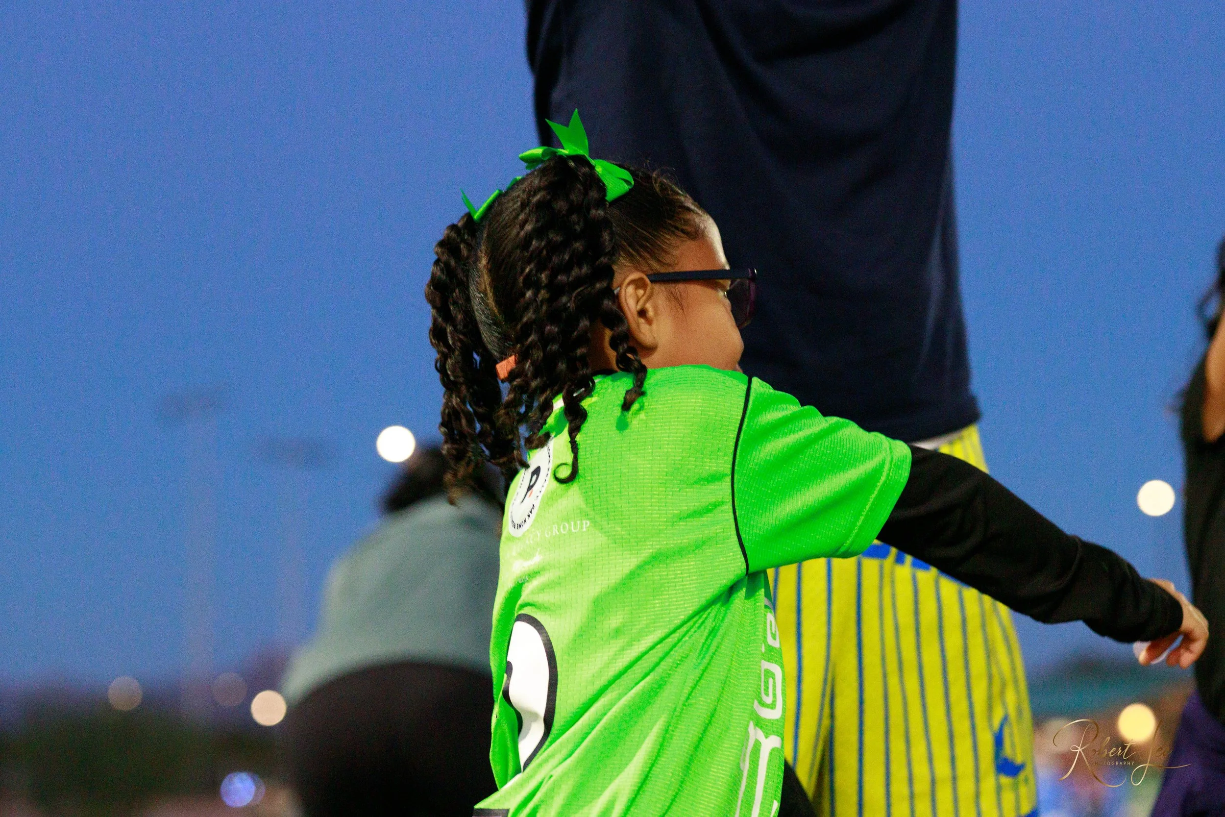 Child with braided hair and glasses wearing a bright green sports jersey, standing outdoors at dusk.