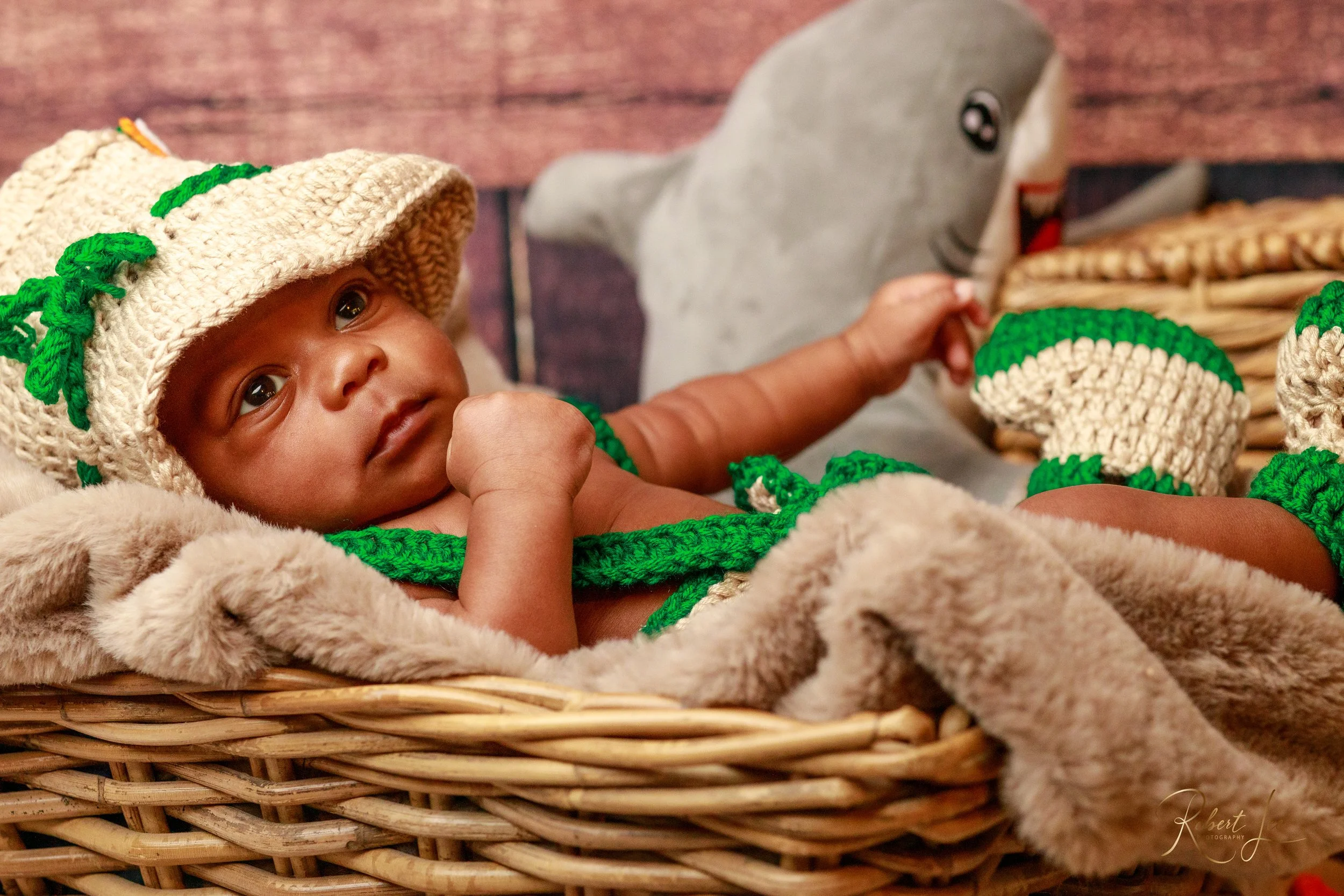 Close-up of a baby lying in a wicker basket, wearing a crochet hat and matching outfit with green accents, holding a stuffed shark toy.