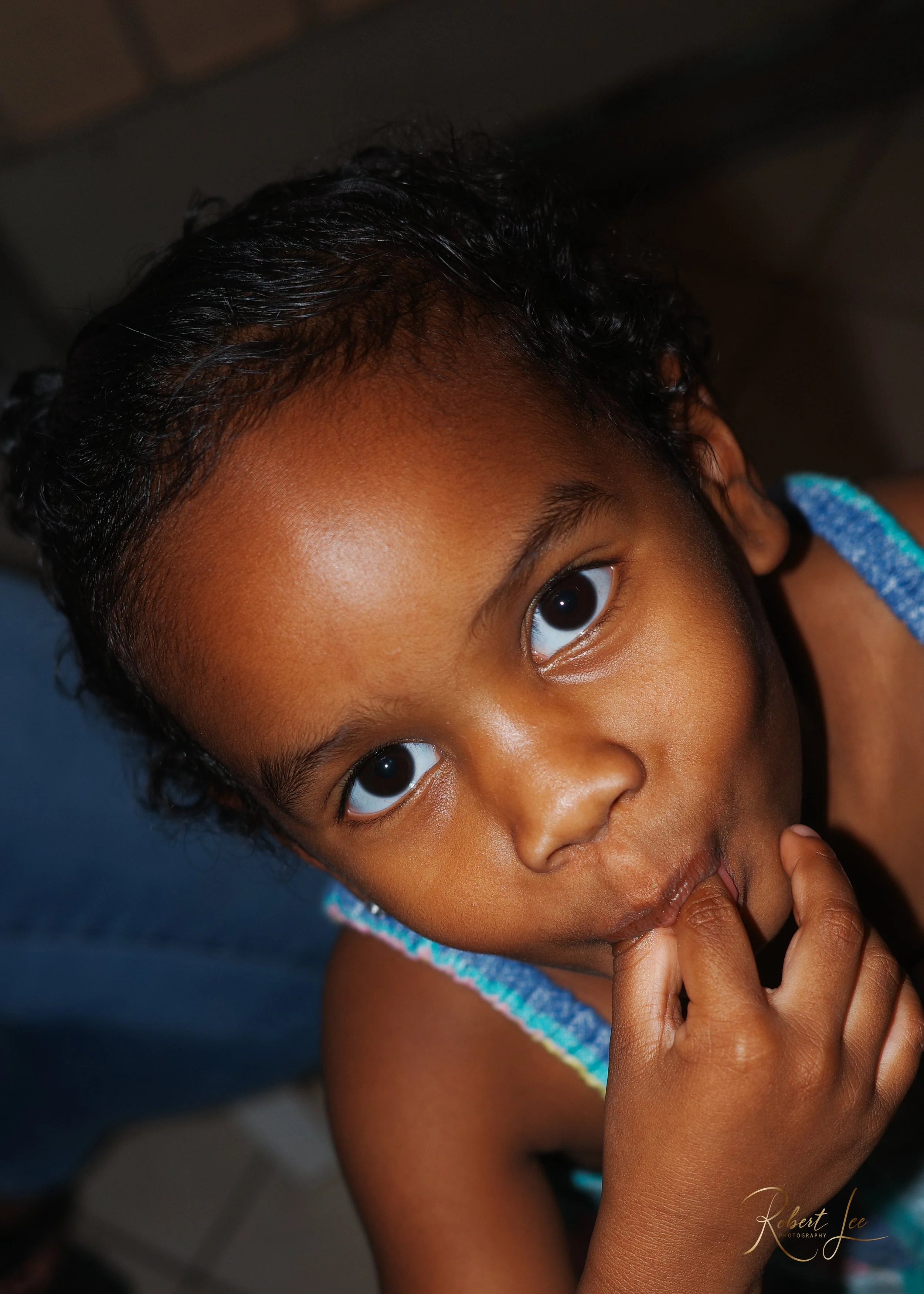 Close-up of a young girl with dark skin, large brown eyes, and curly hair, looking into the camera with her hand near her mouth.