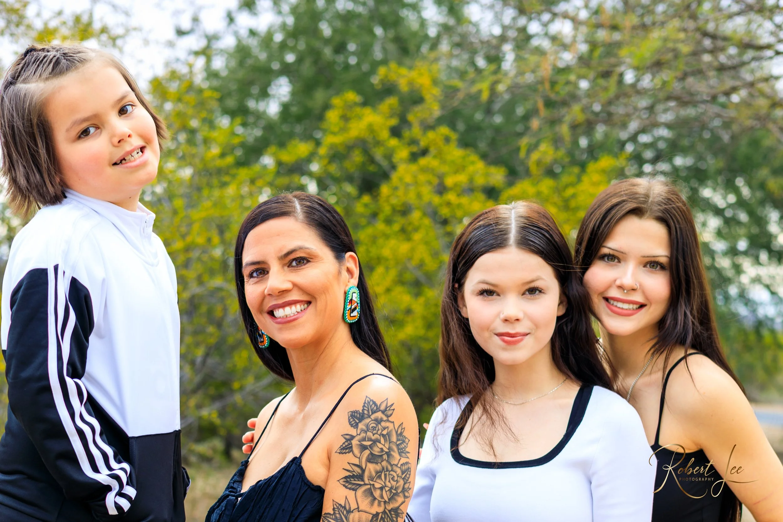 Four women and one young girl outdoors on a cloudy day, with trees in the background. They are smiling and looking at the camera, wearing casual clothing.