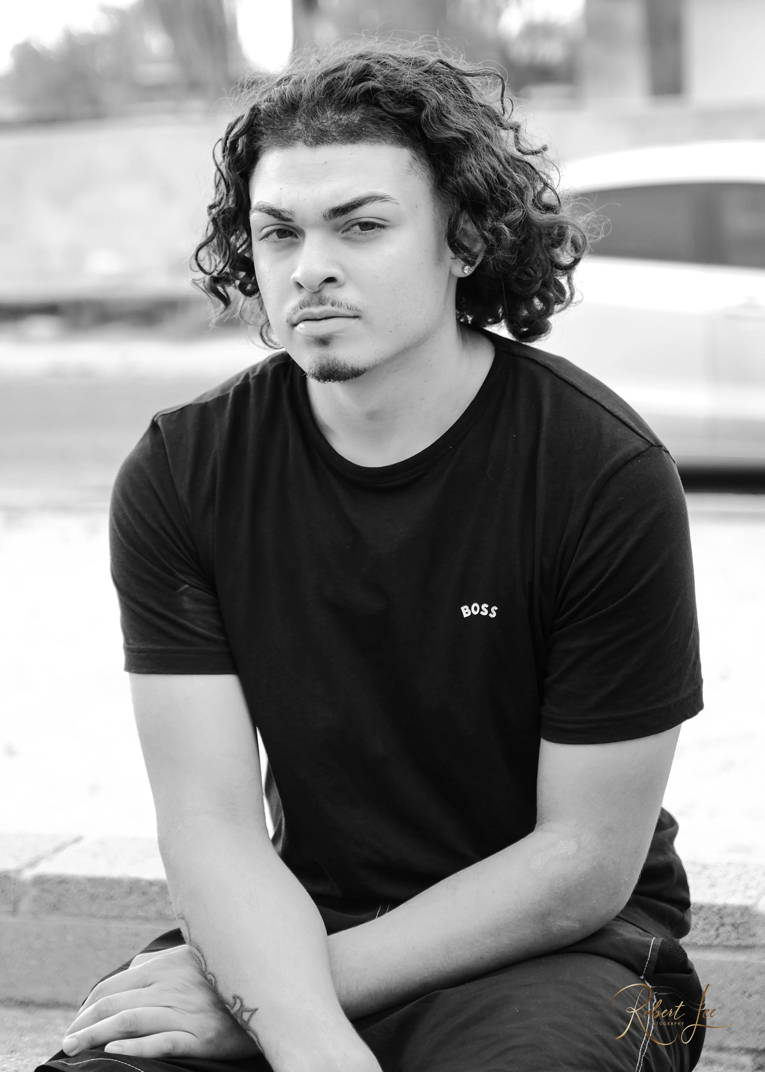 Black and white portrait of a young man with curly hair, wearing a black t-shirt with 'BOSS' written on it, sitting outdoors.