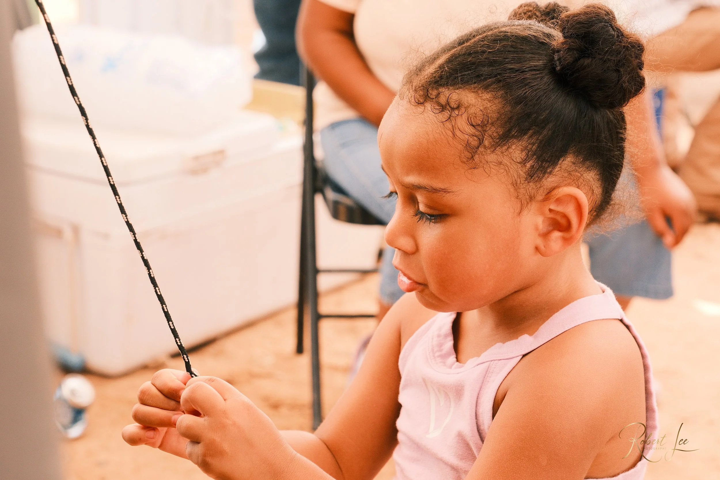 Young girl with curly hair in a bun looks down at a string she is holding in both hands, sitting indoors around other people.