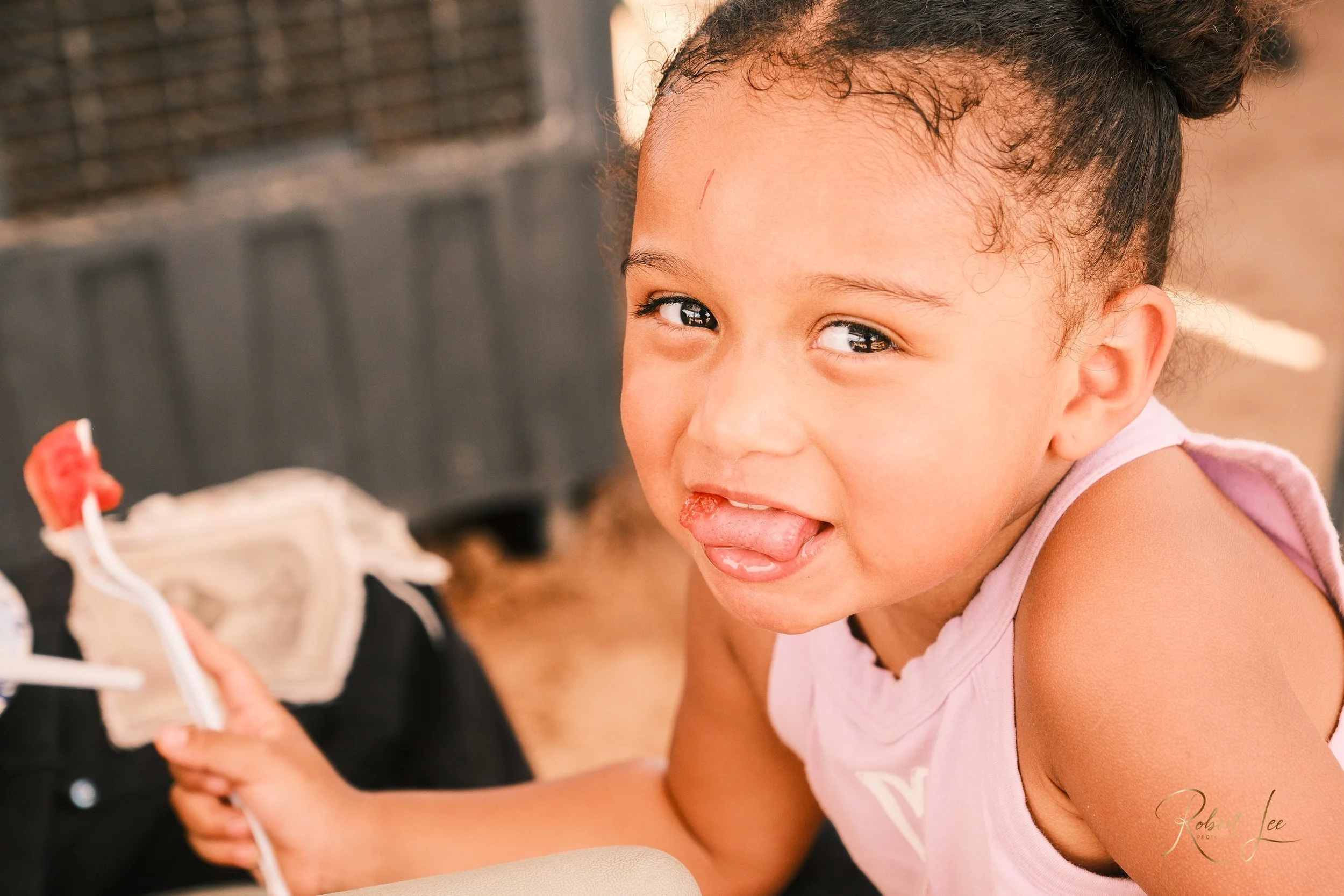 A young girl with curly hair tied in a bun, sticking out her tongue and smiling, holding a fork.