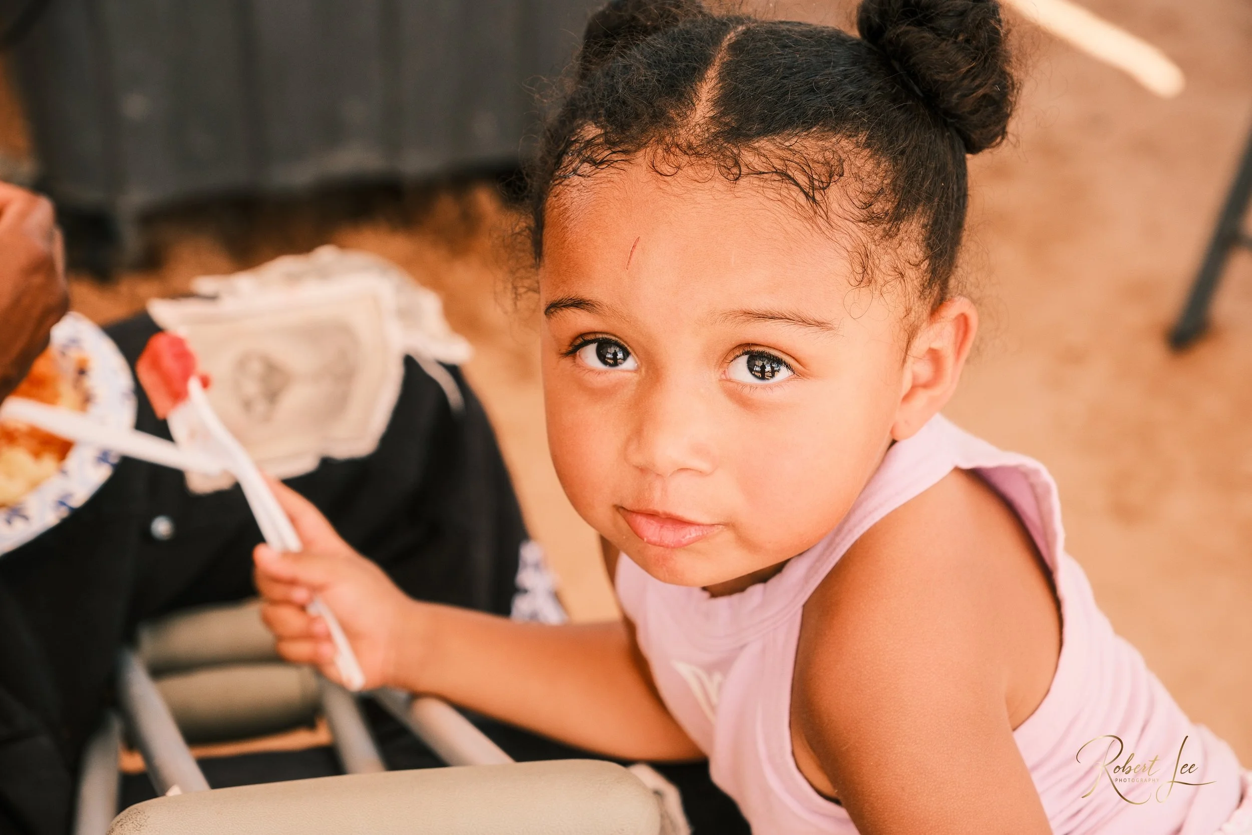 A young girl with curly hair in a bun holding a plush toy, looking up at the camera with big eyes, wearing a pink sleeveless top.