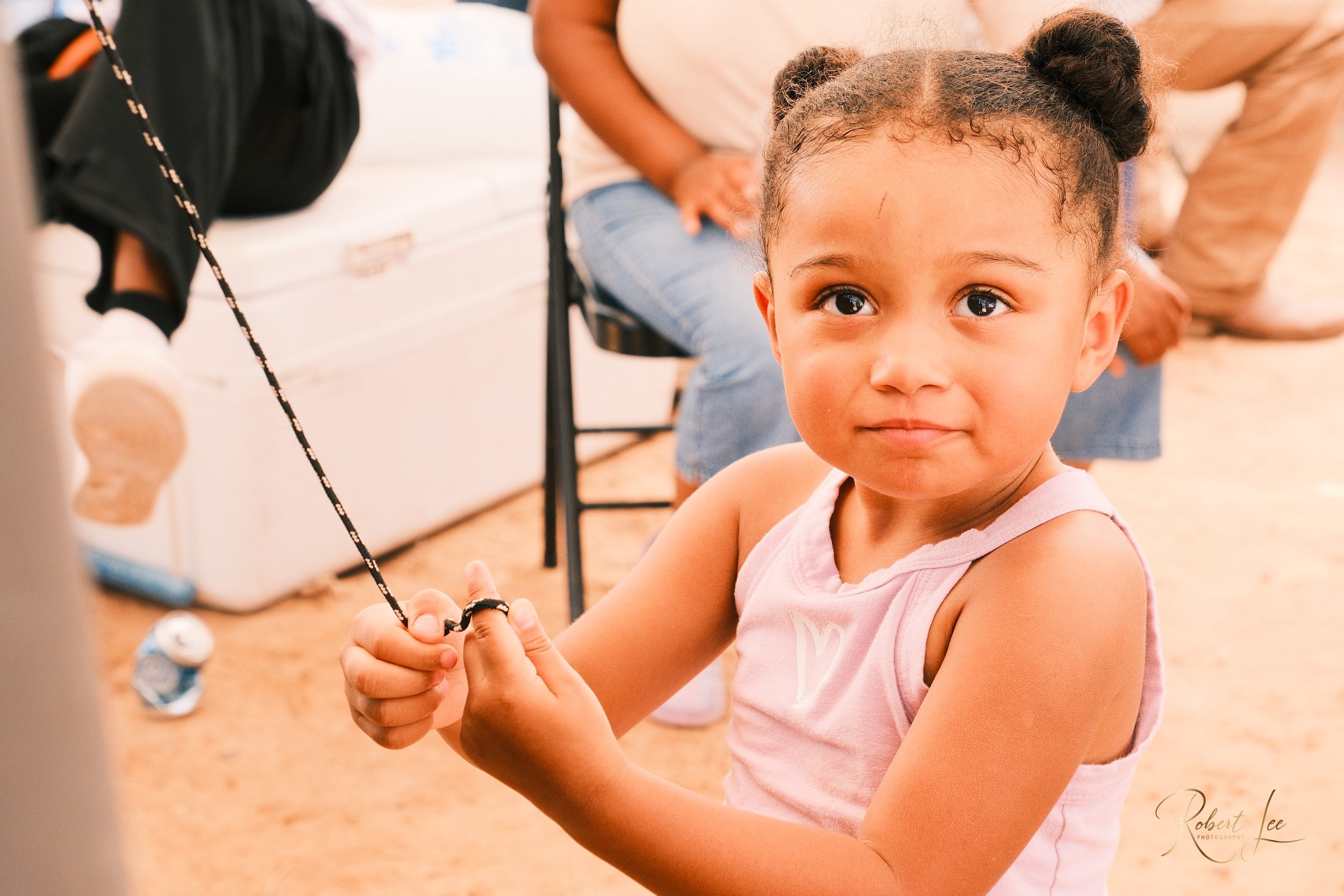 Young girl with braided hair and a pink tank top looking at the camera while holding a string or cord, sitting indoors.