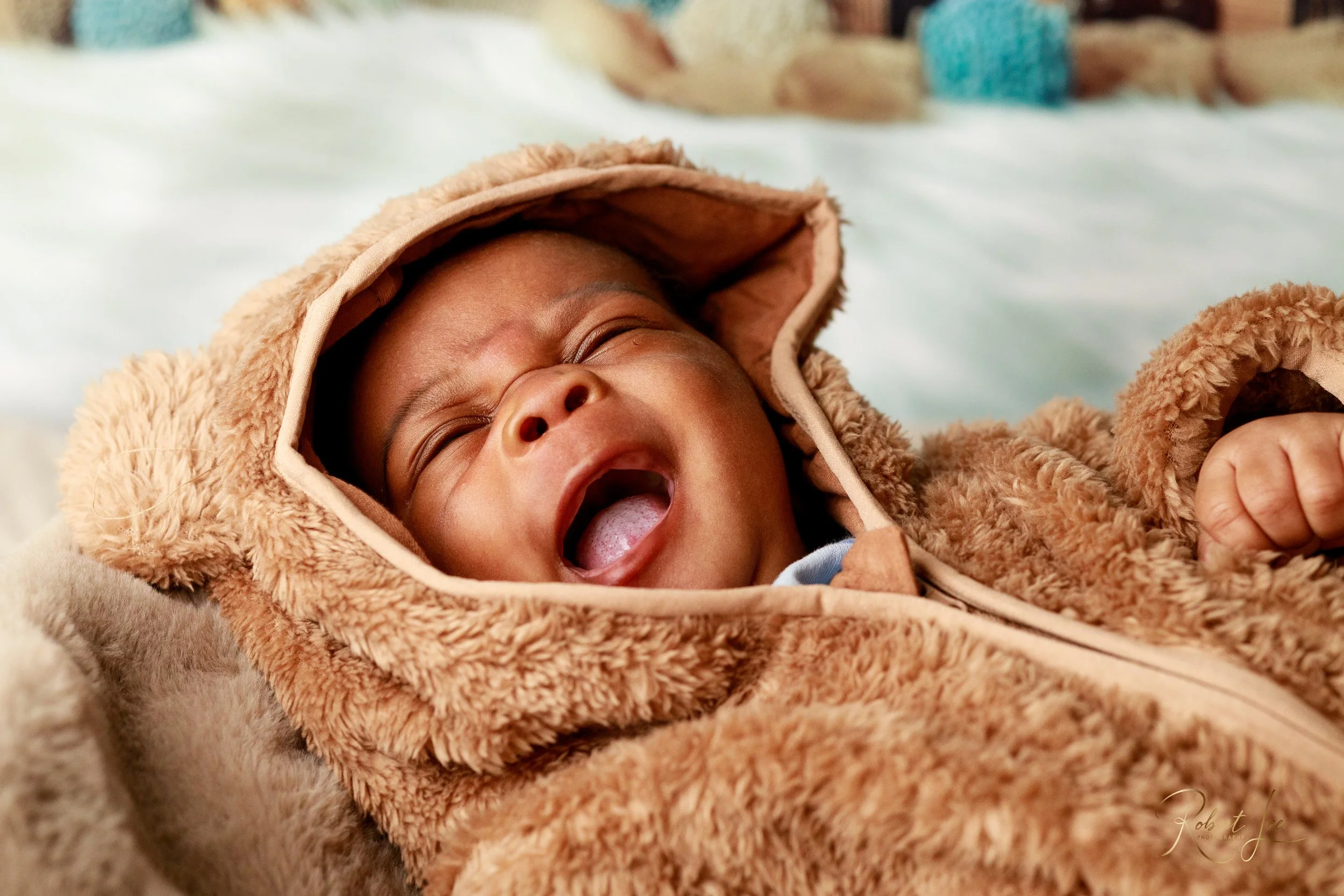 A baby yawning while lying on a soft surface, wearing a cozy brown hooded jacket with furry lining.