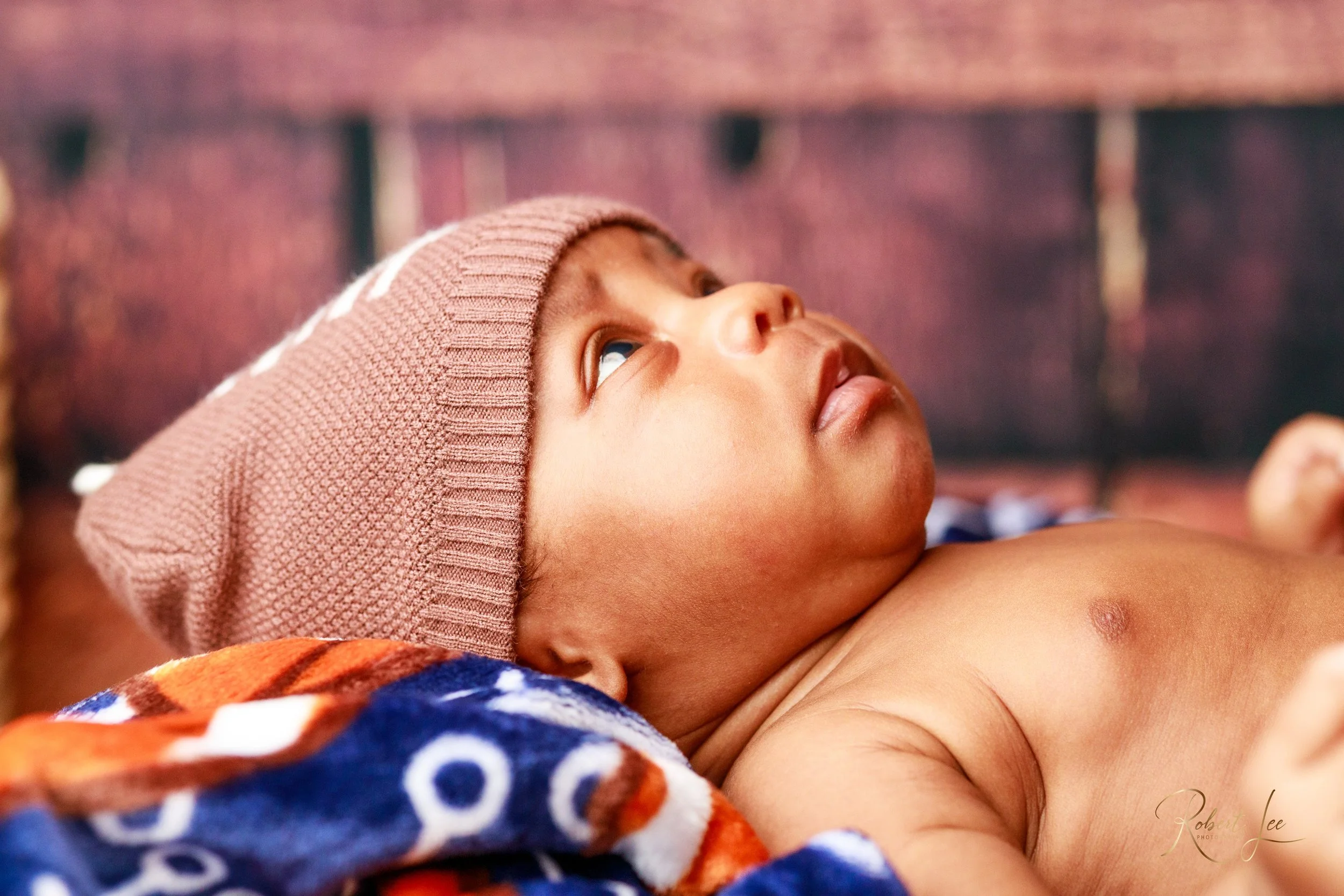 A young child lying on a colorful blanket, wearing a knit cap, looking upward with a curious expression.
