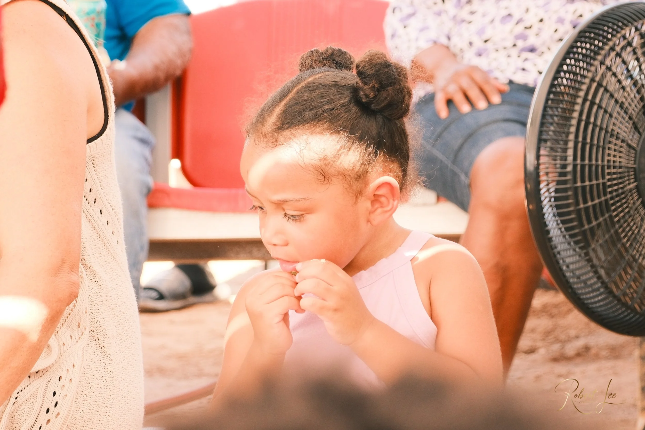Young girl with hair in buns eating a snack, with people sitting nearby and a fan in the foreground.