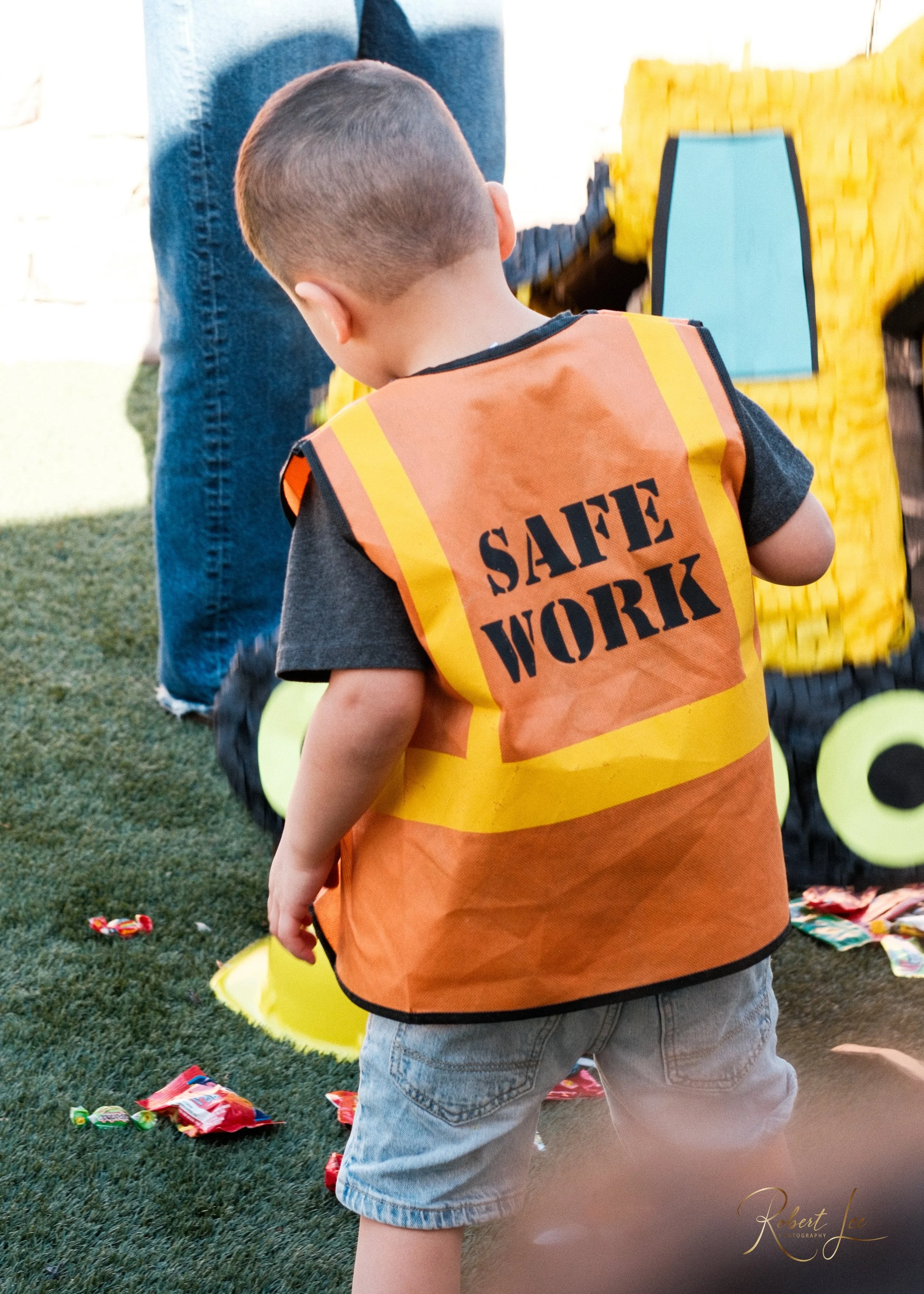 A young boy wearing an orange safety vest labeled 'SAFE WORK' is playing outside near a toy truck and scattered candy on the grass.