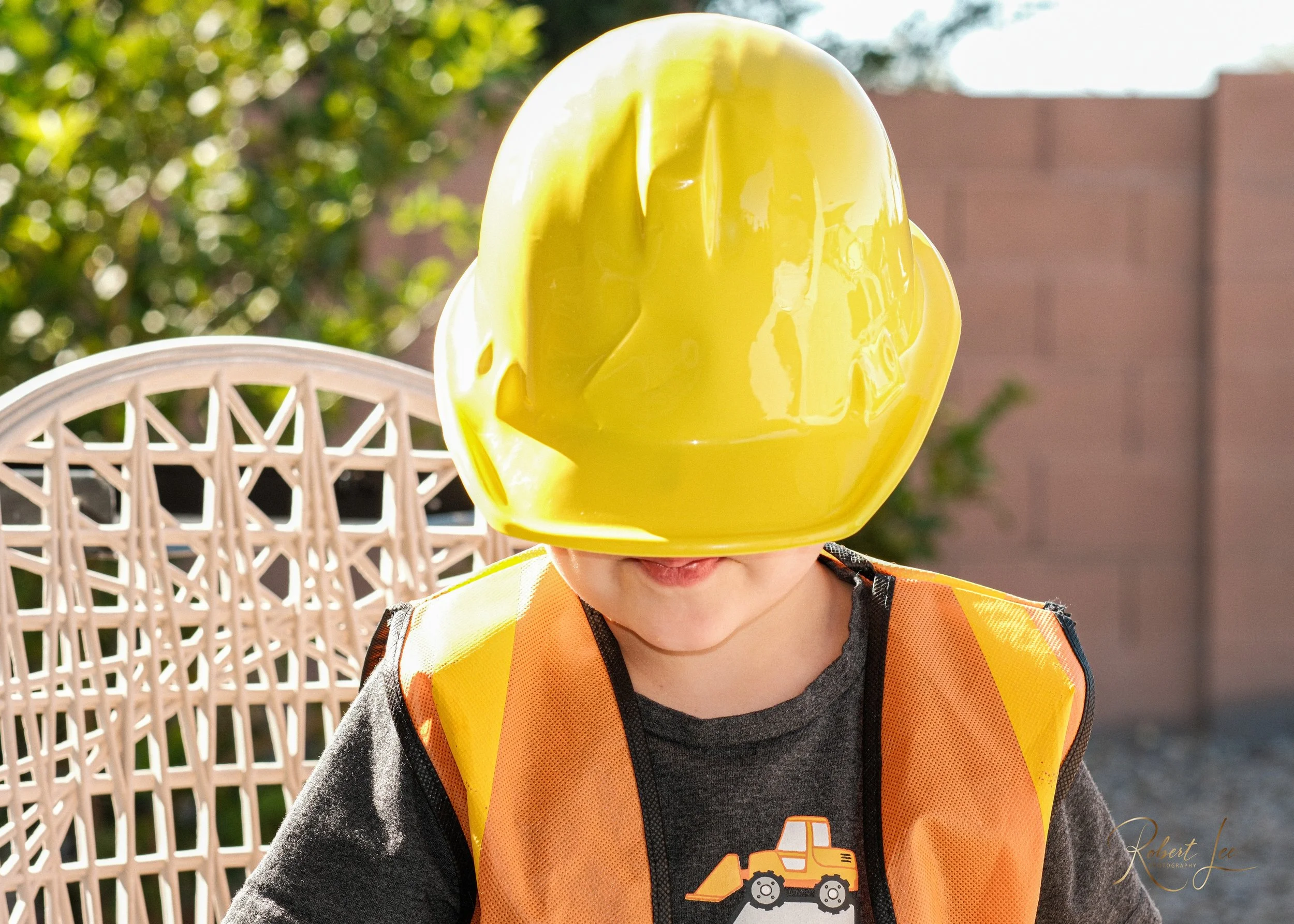 Child wearing a yellow construction helmet and orange safety vest, sitting outdoors on a white lattice chair, with trees and a brick wall in the background.