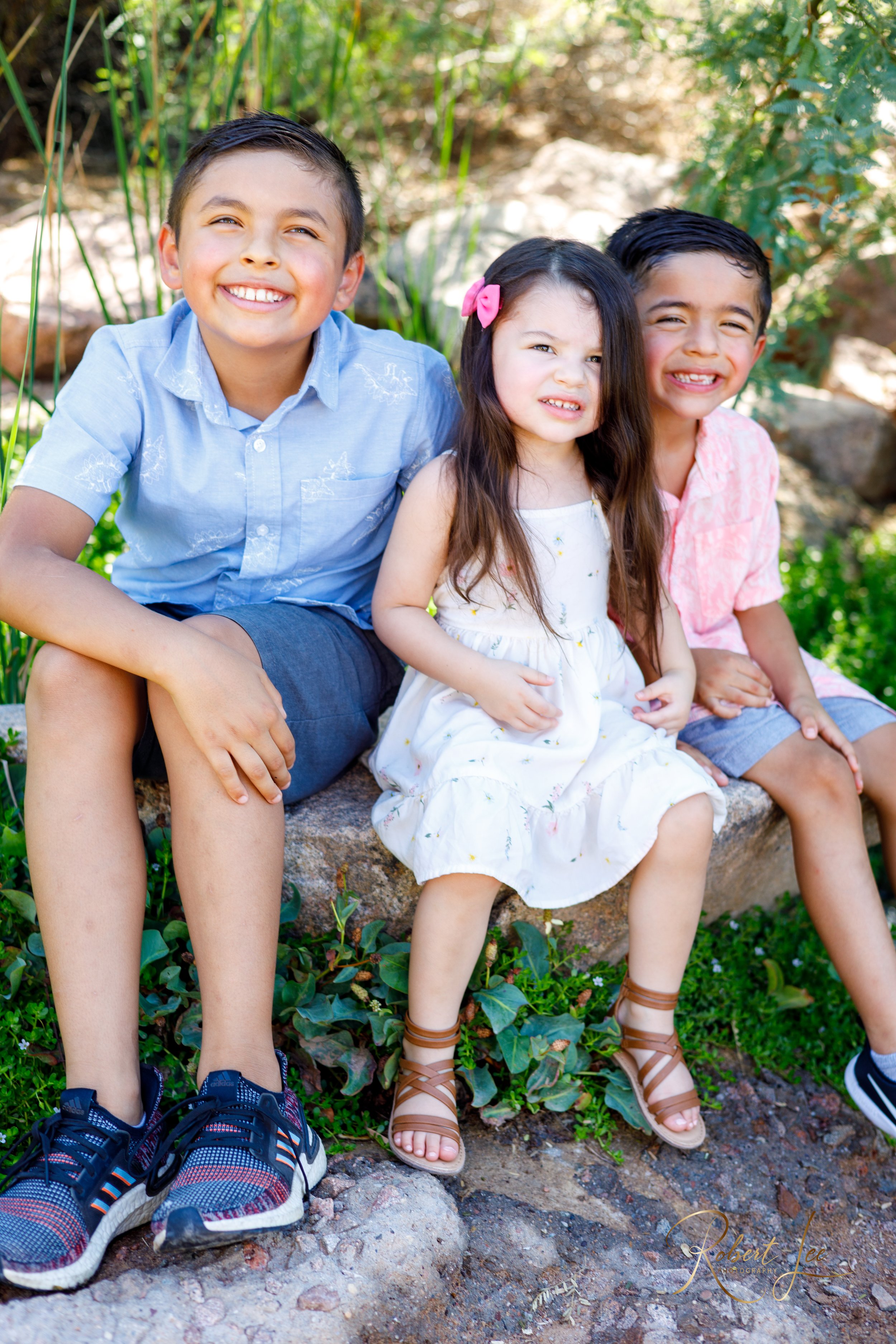 Three children sitting on a stone bench outside surrounded by greenery, smiling and making funny faces. Tucson Portrait photographer. Robert lee Photography.