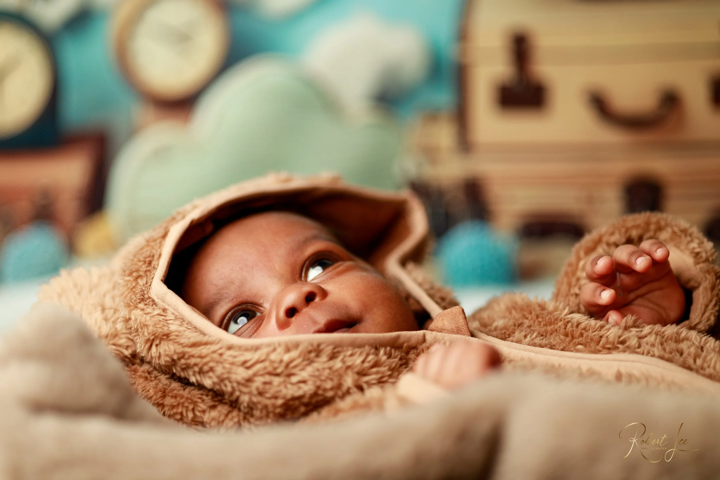 A baby lying down wearing a brown fuzzy hooded jacket, looking upwards with wide eyes in a cozy, colorful setting.