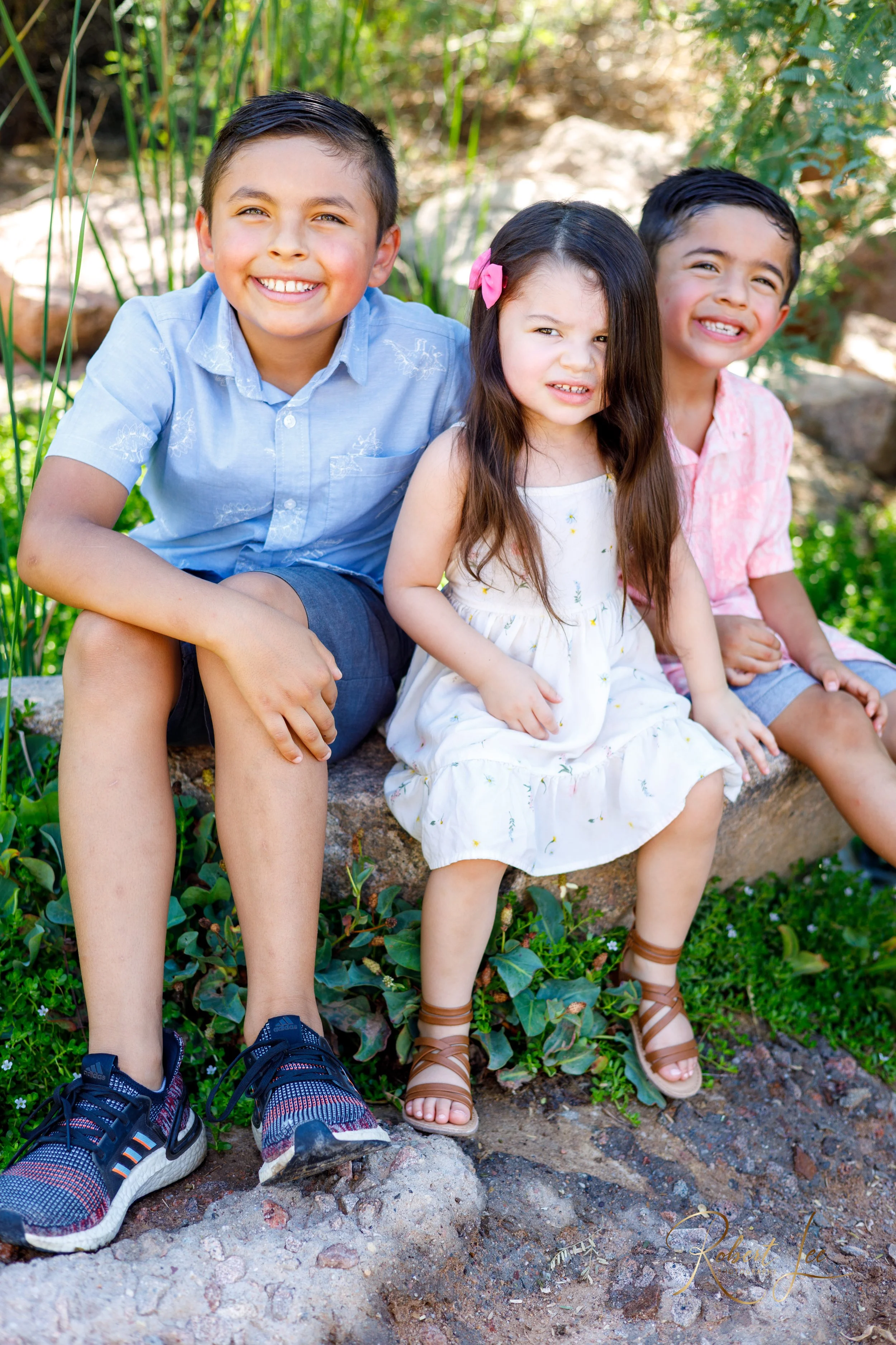 Three children sitting outdoors on a stone edge with greenery in the background. The girl in the middle has long brown hair with a pink bow, wearing a white dress, and makes a silly face. The boy on the right is smiling, wearing a pink shirt and shor
