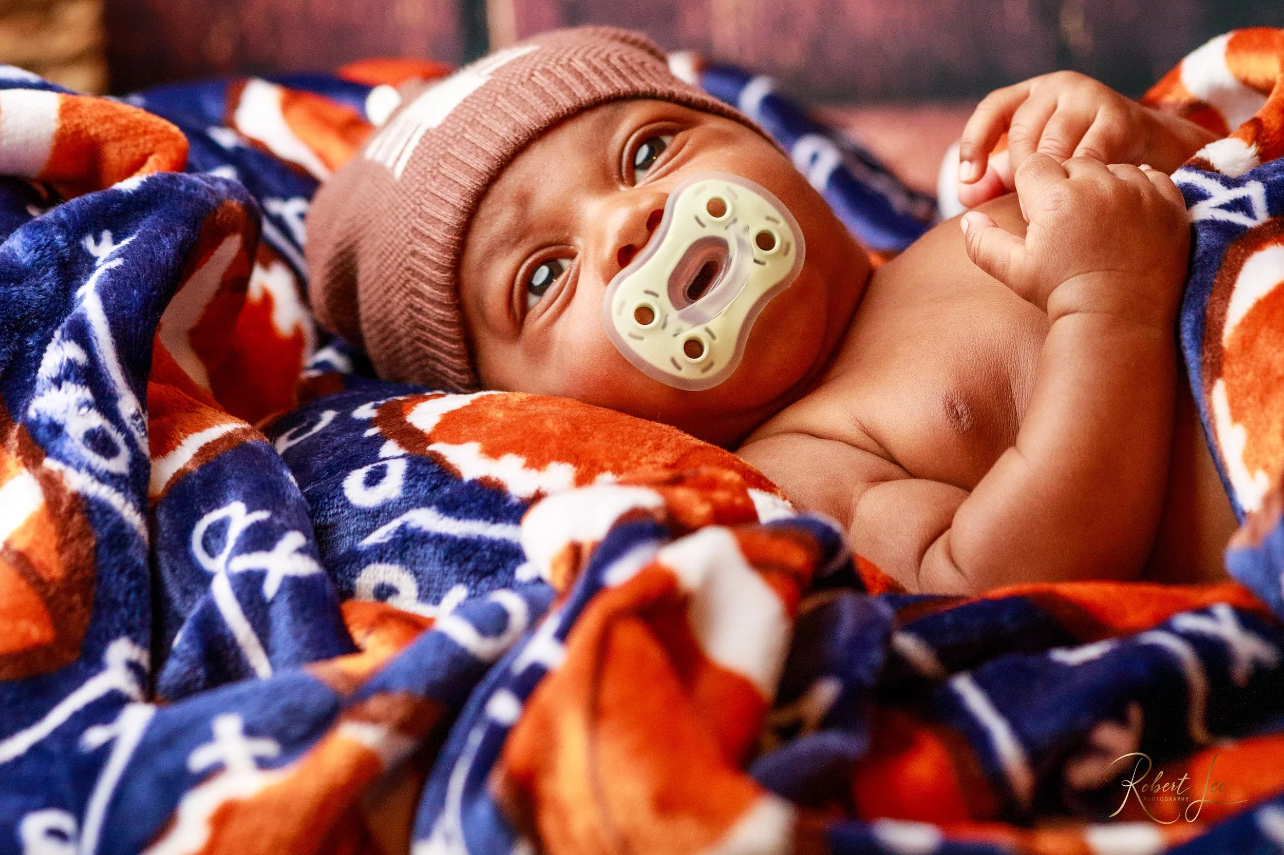 Close-up of a baby lying on a colorful blanket, wearing a brown beanie and with a pacifier in their mouth, looking up.