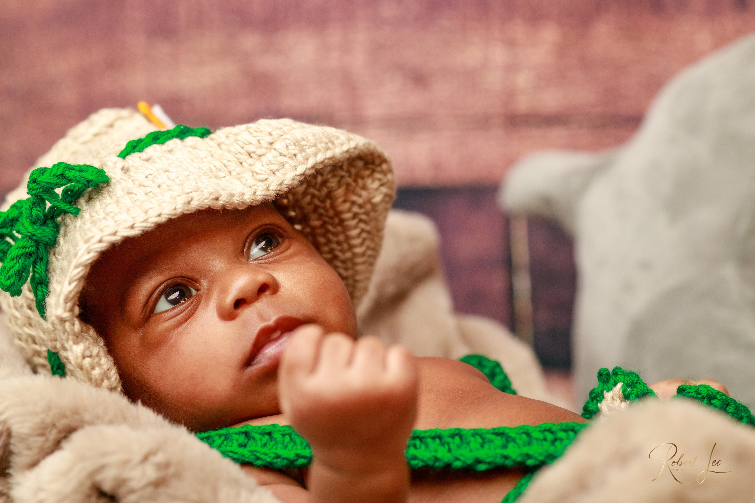 Close-up of a baby wearing a beige knitted hat with green details, lying on a soft surface and looking up with curiosity.