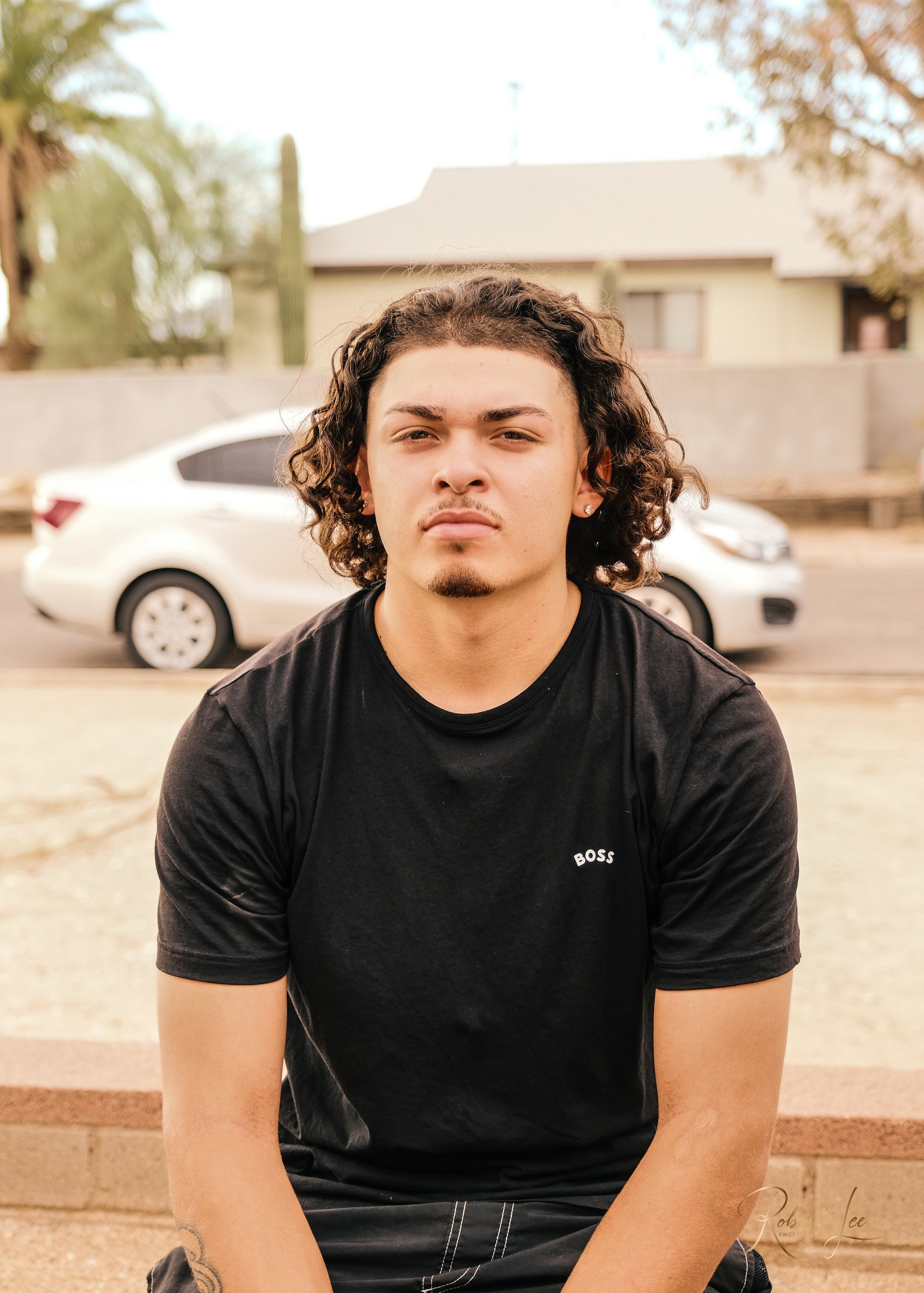 Young man with curly hair and a goatee sitting outdoors in front of a white car, wearing a black T-shirt with BOSS on it.