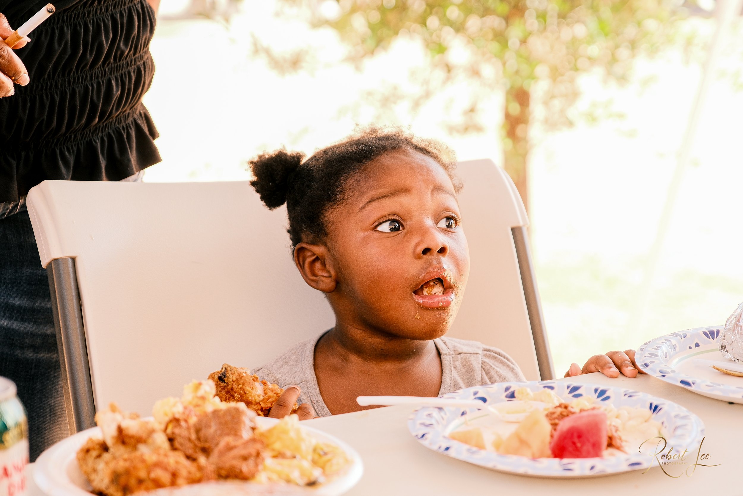 A young girl with curly hair in pigtails sitting at a table with plates of food, looking surprised or shocked, with food on her face.