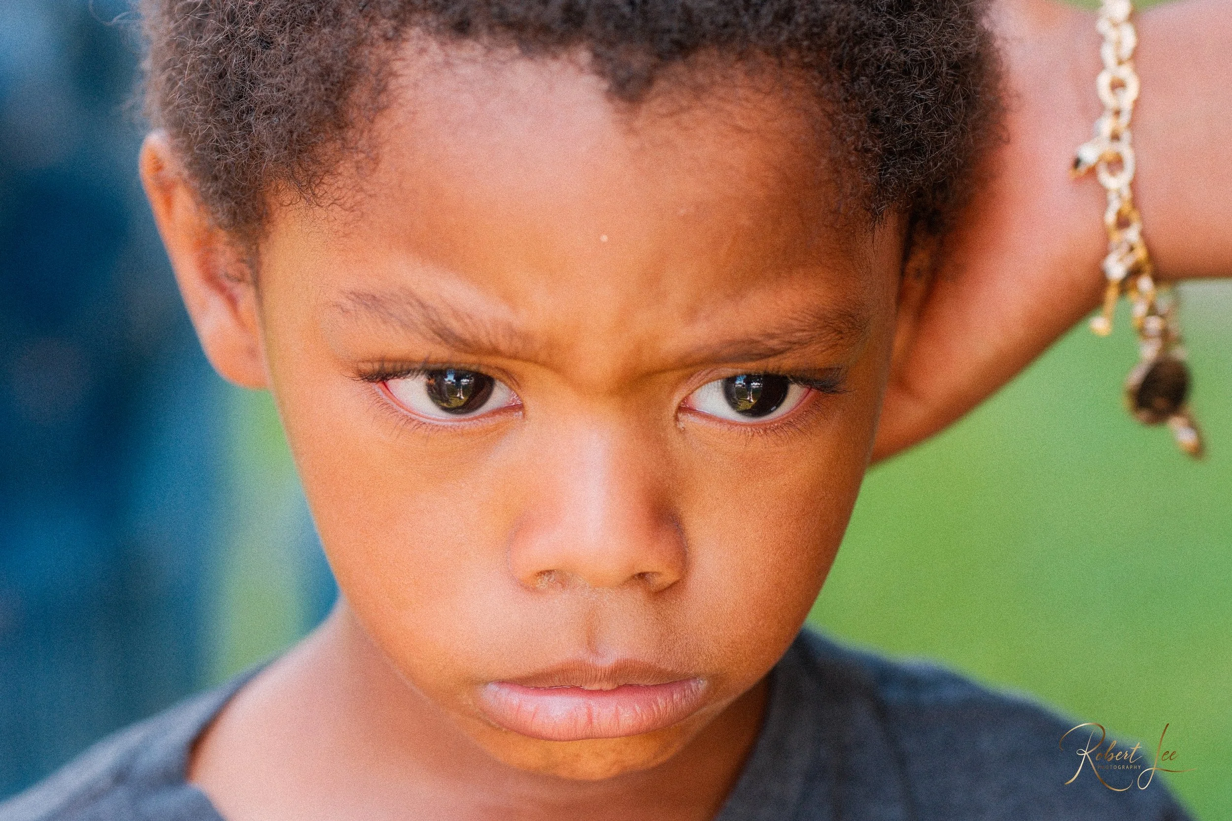 A young child with a serious expression, looking downward with intense eyes, wearing a black shirt and a gold bracelet, with a blurred outdoor background.
