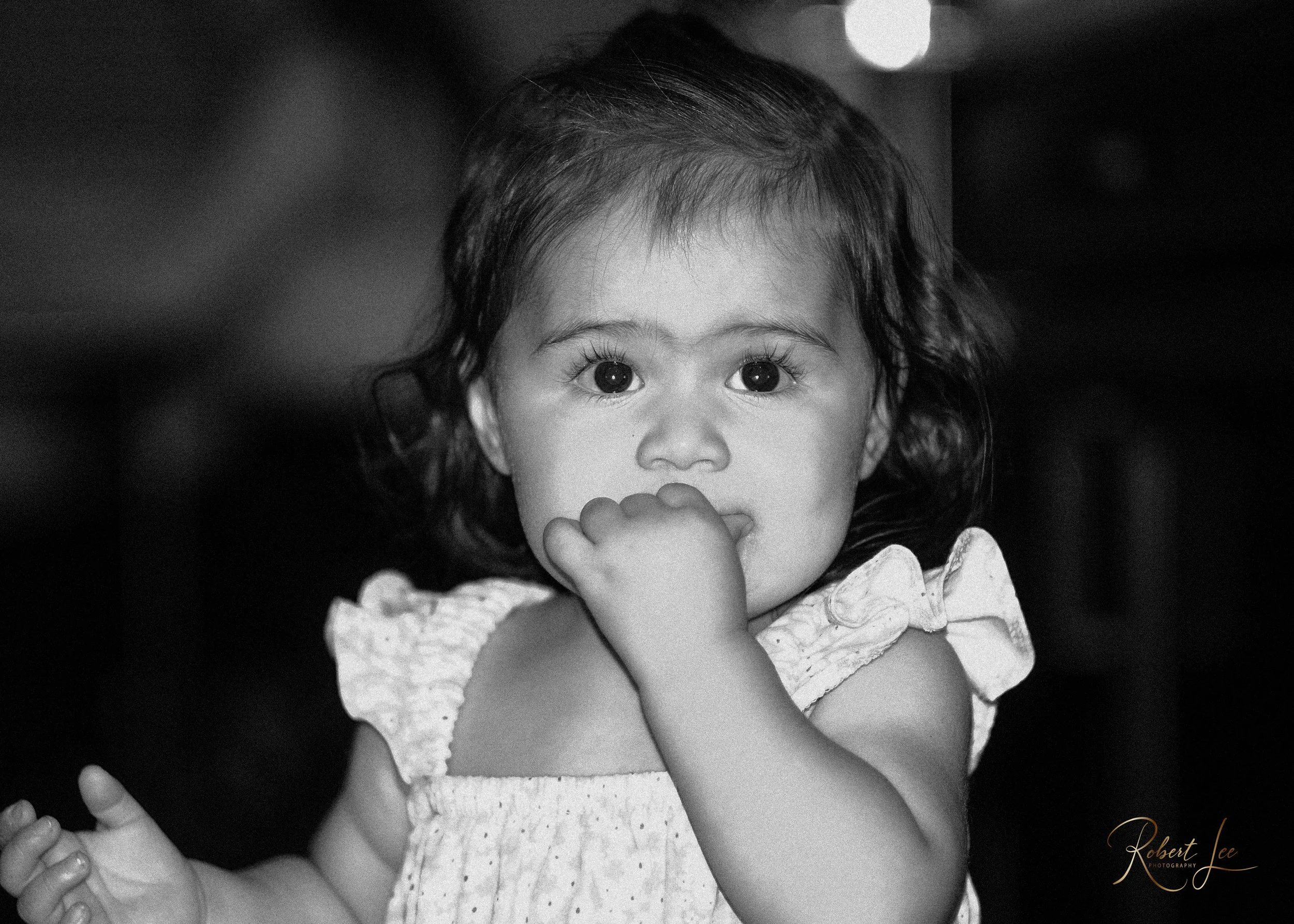 A young girl with large, expressive eyes and dark hair, wearing a ruffled sleeveless top, is seen touching her lips with her right hand, looking directly at the camera. The photo is in black and white.