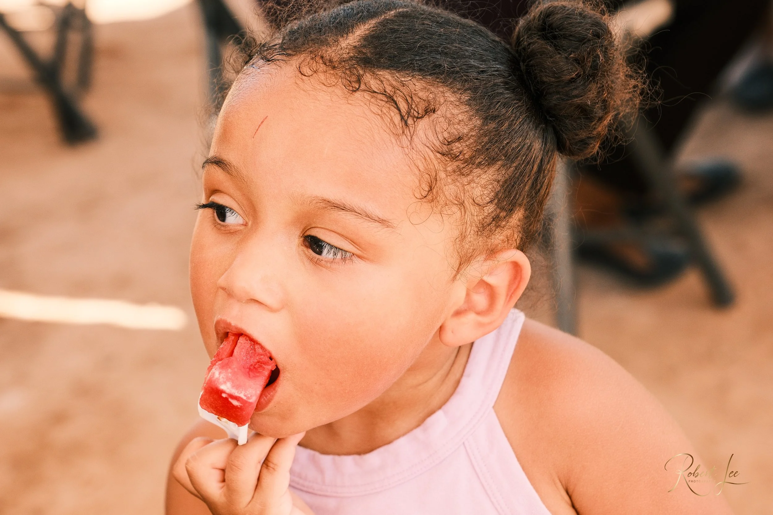 A young girl with braided hair in buns, wearing a light pink top, eating a red popsicle with white streaks.