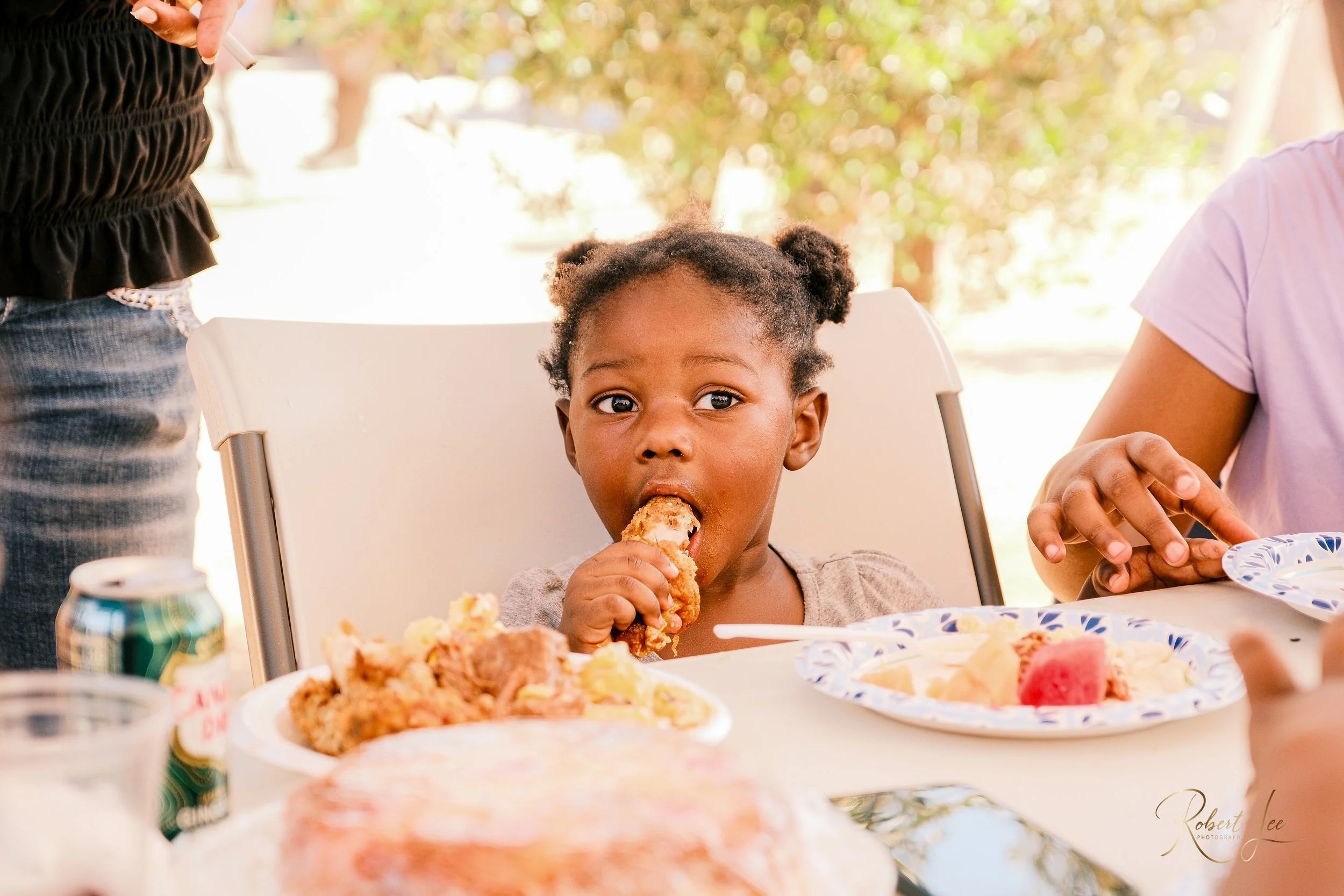 A young girl with dark hair in puffs sitting at a table and eating fried chicken, with plates of food including watermelon, and a can of soda in front of her. The setting appears to be outdoors.