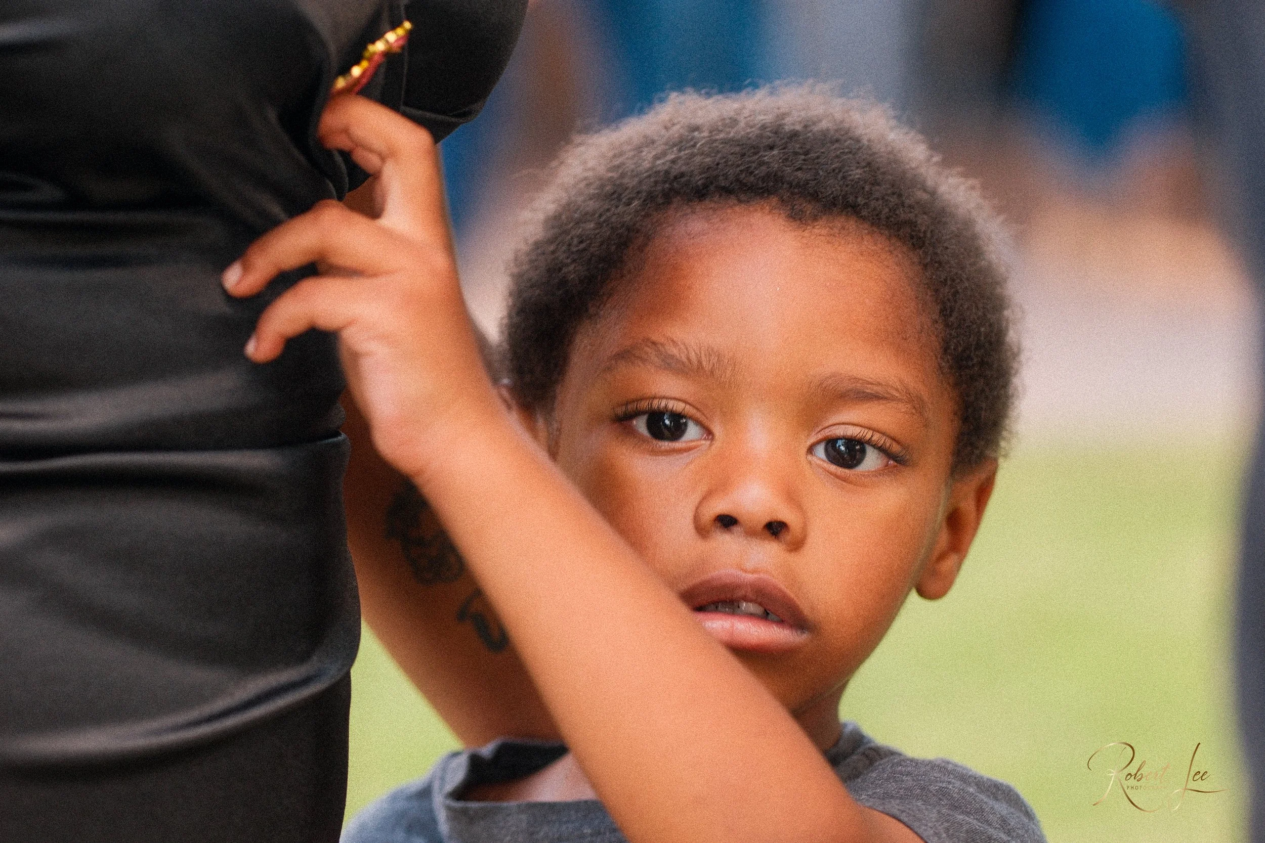 A young boy looking with curiosity and slight concern, with an adult, partially visible, standing nearby in an outdoor setting.
