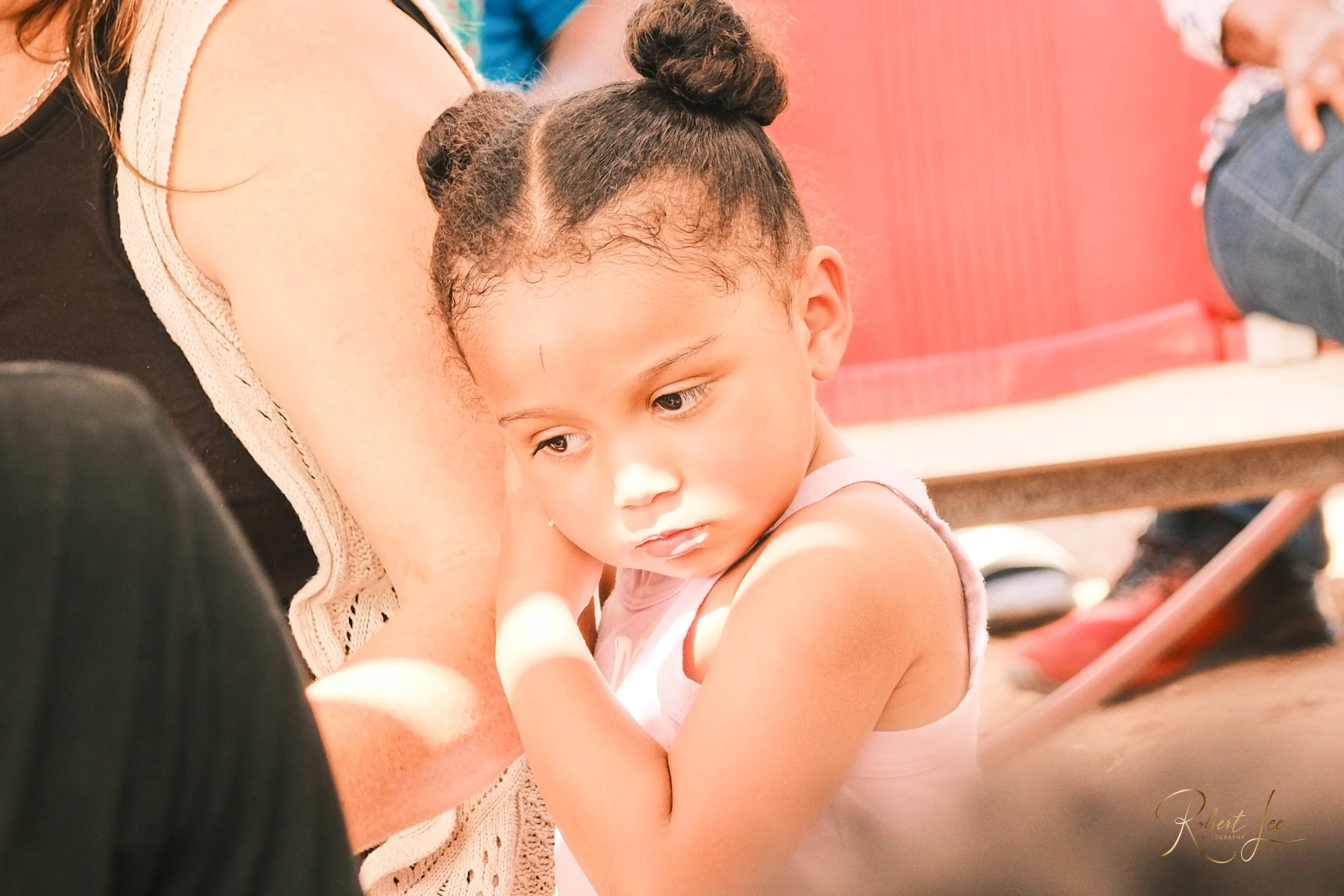 A young girl with pigtails resting her head on an adult's shoulder, looking down with a thoughtful expression.