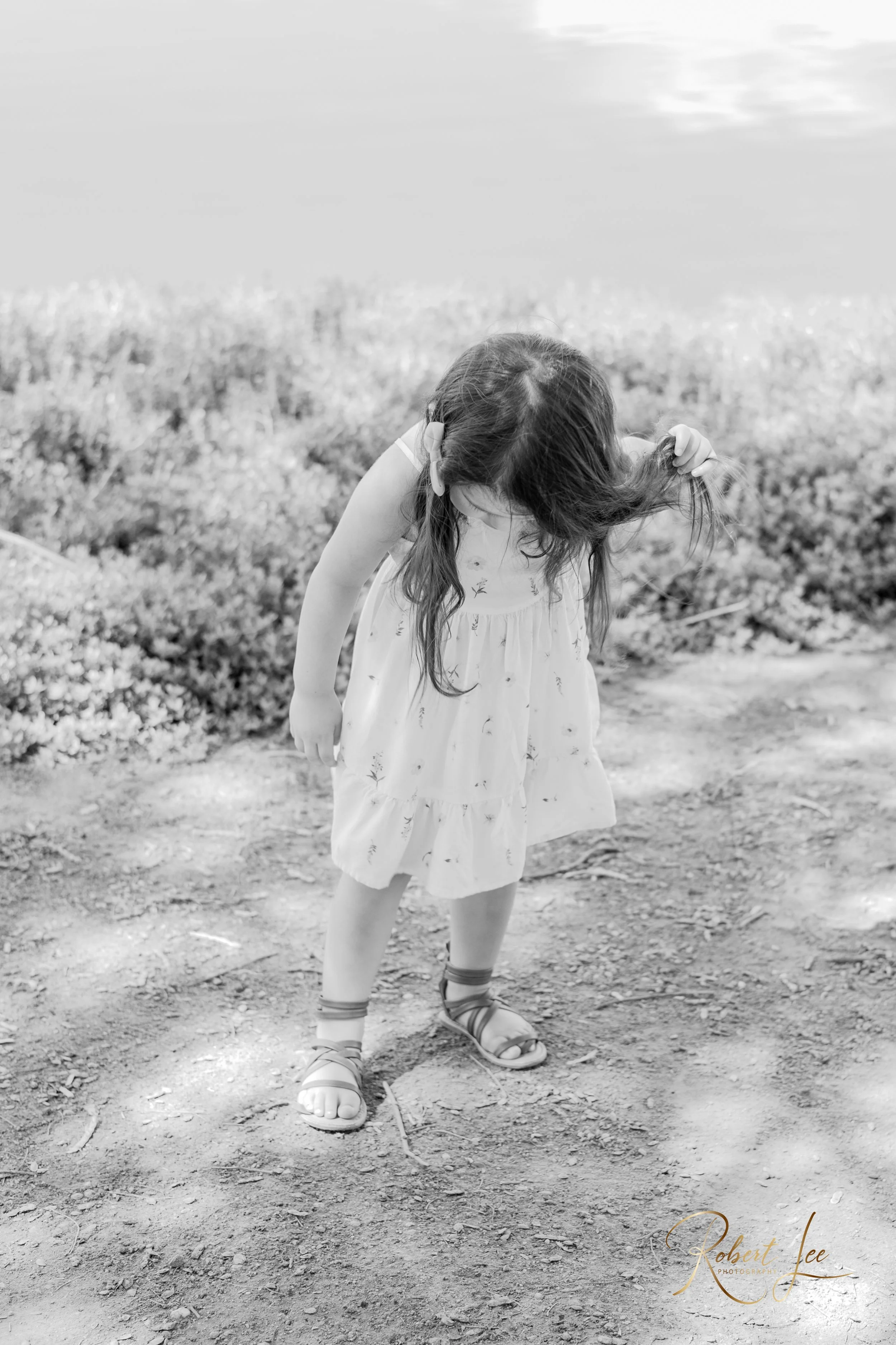 A young girl with long hair, wearing a sleeveless dress and sandals, is standing on a dirt path outdoors, bending down while holding her hair with her right hand. There are bushes and a body of water in the background. Tucson Portrait photographer. R