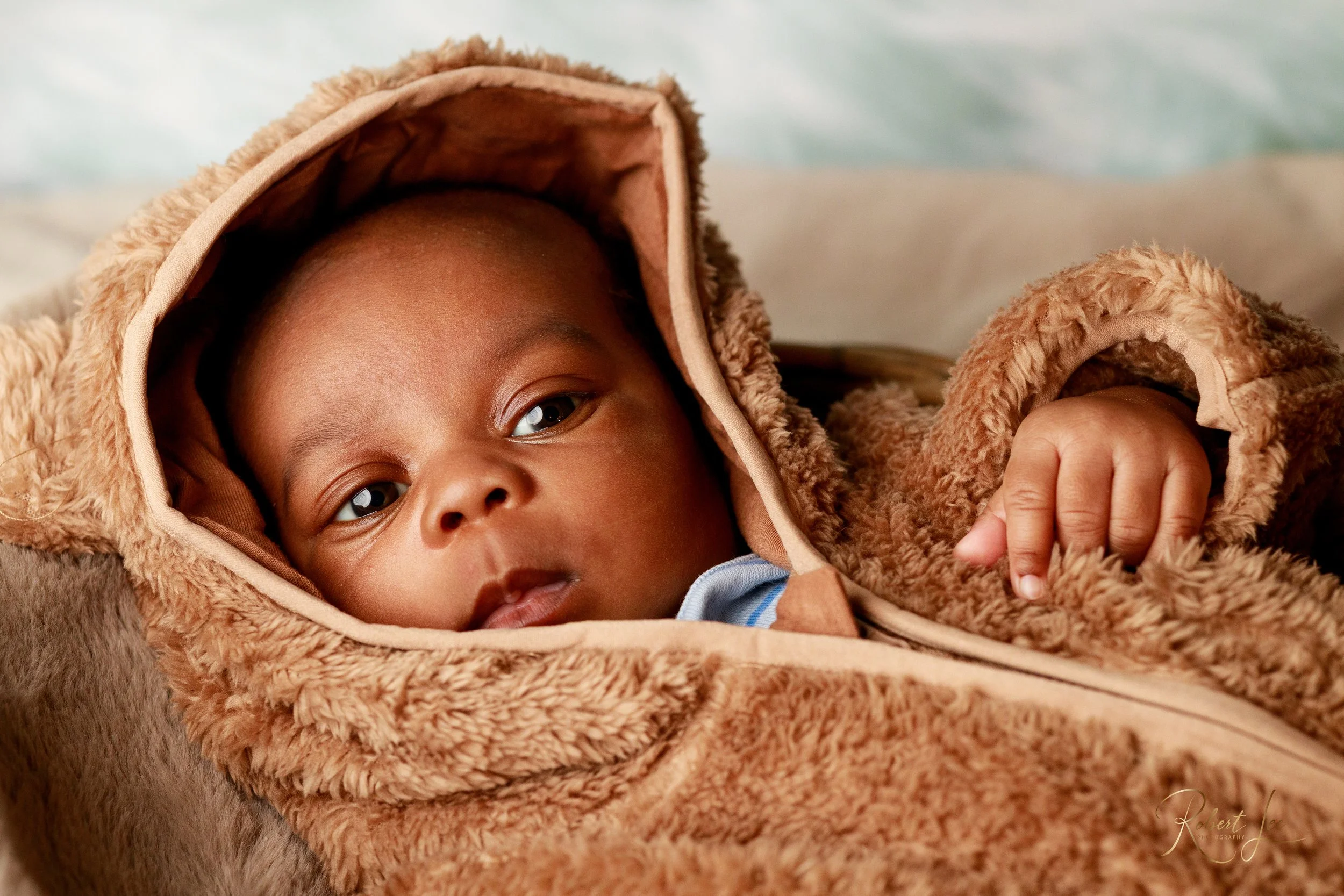 A baby lying in a fuzzy brown hoodie with bear ears, looking directly at the camera.