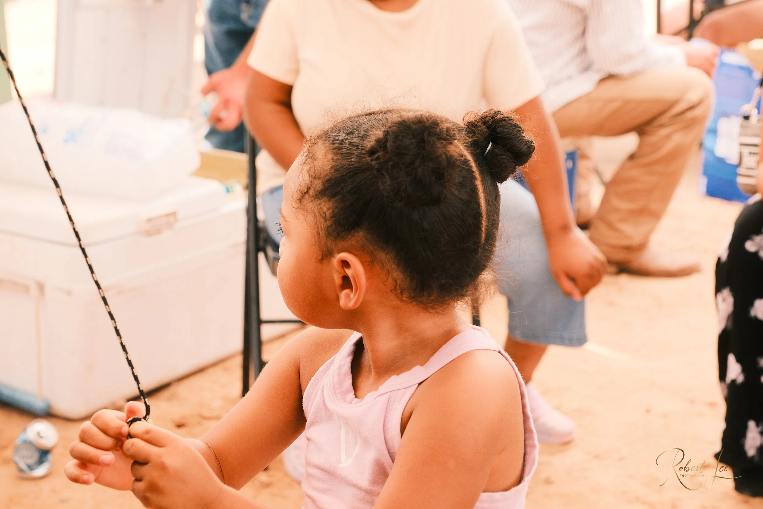 A young girl with black hair in two buns holds a black and white braided string, with adults sitting nearby in a casual indoor setting.