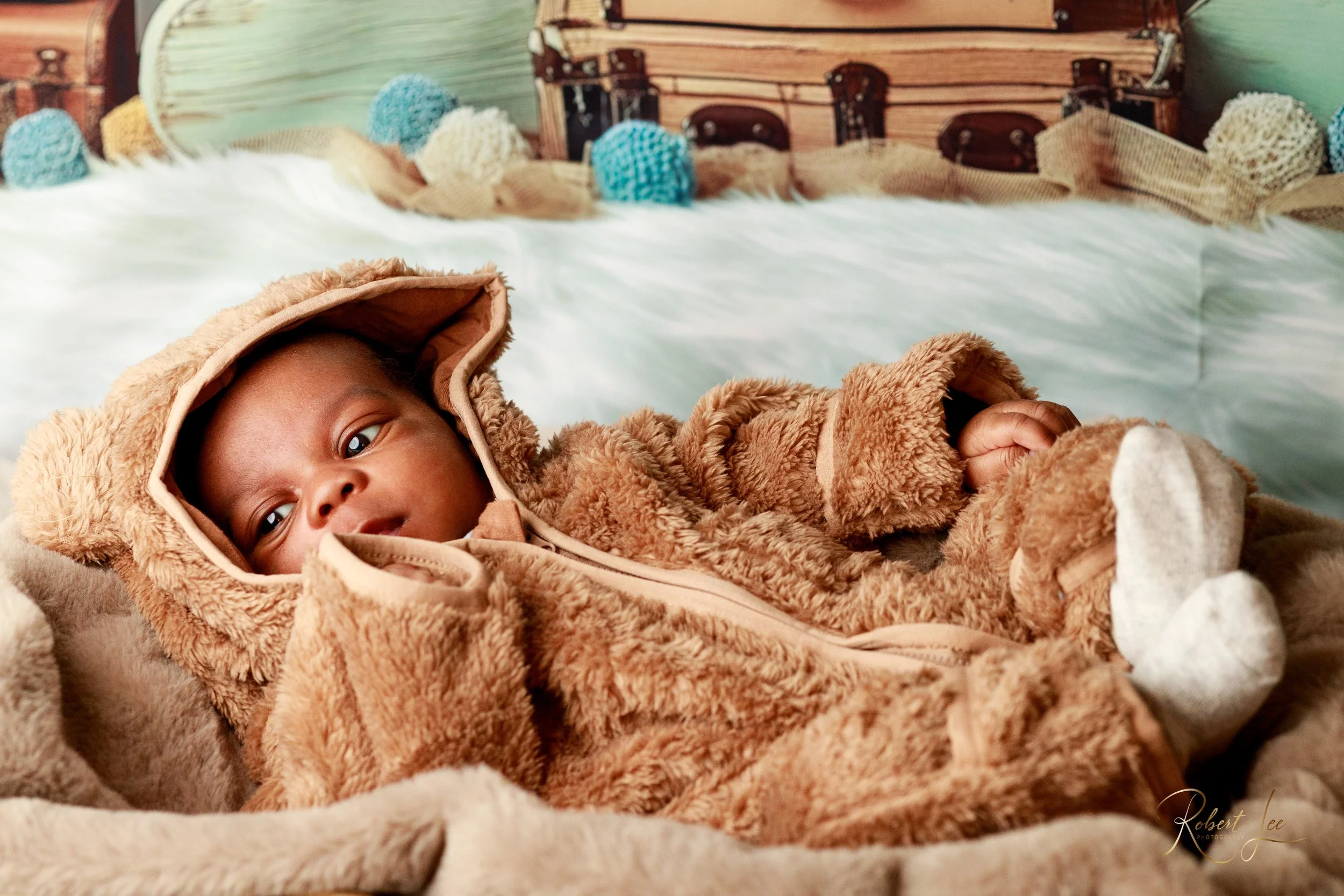 A baby dressed in a brown teddy bear costume lying on a bed with white bedding, surrounded by soft colored yarn pom-poms and woven baskets in the background.