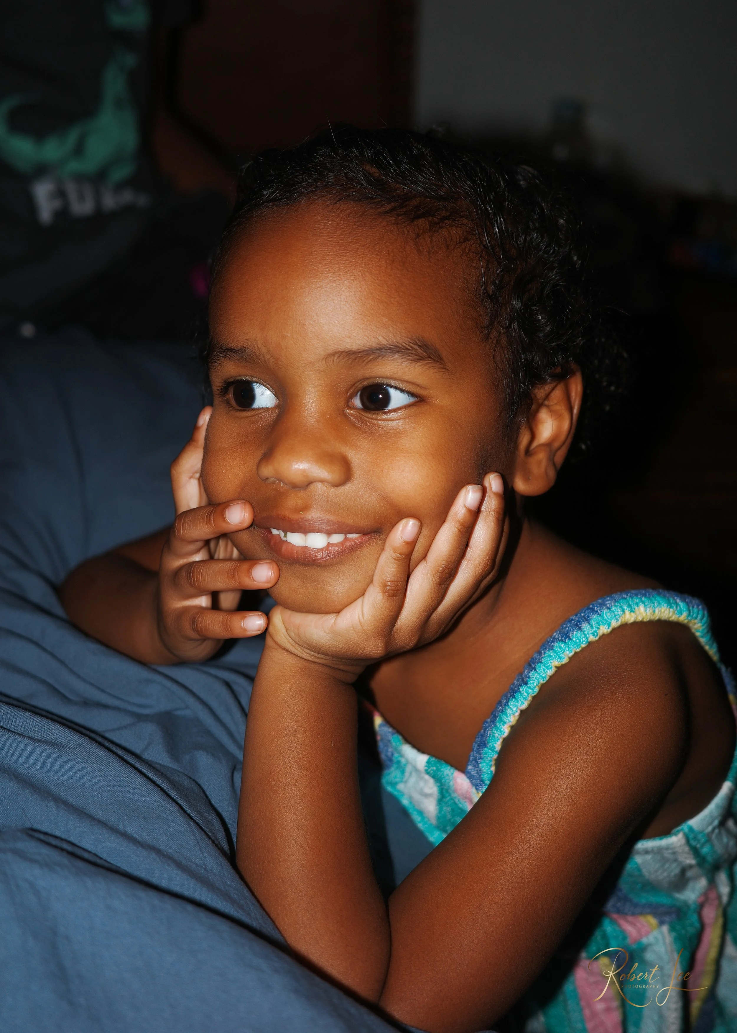 Young girl with brown skin and dark curly hair smiling, resting her chin on her hands with elbows on a blue surface.