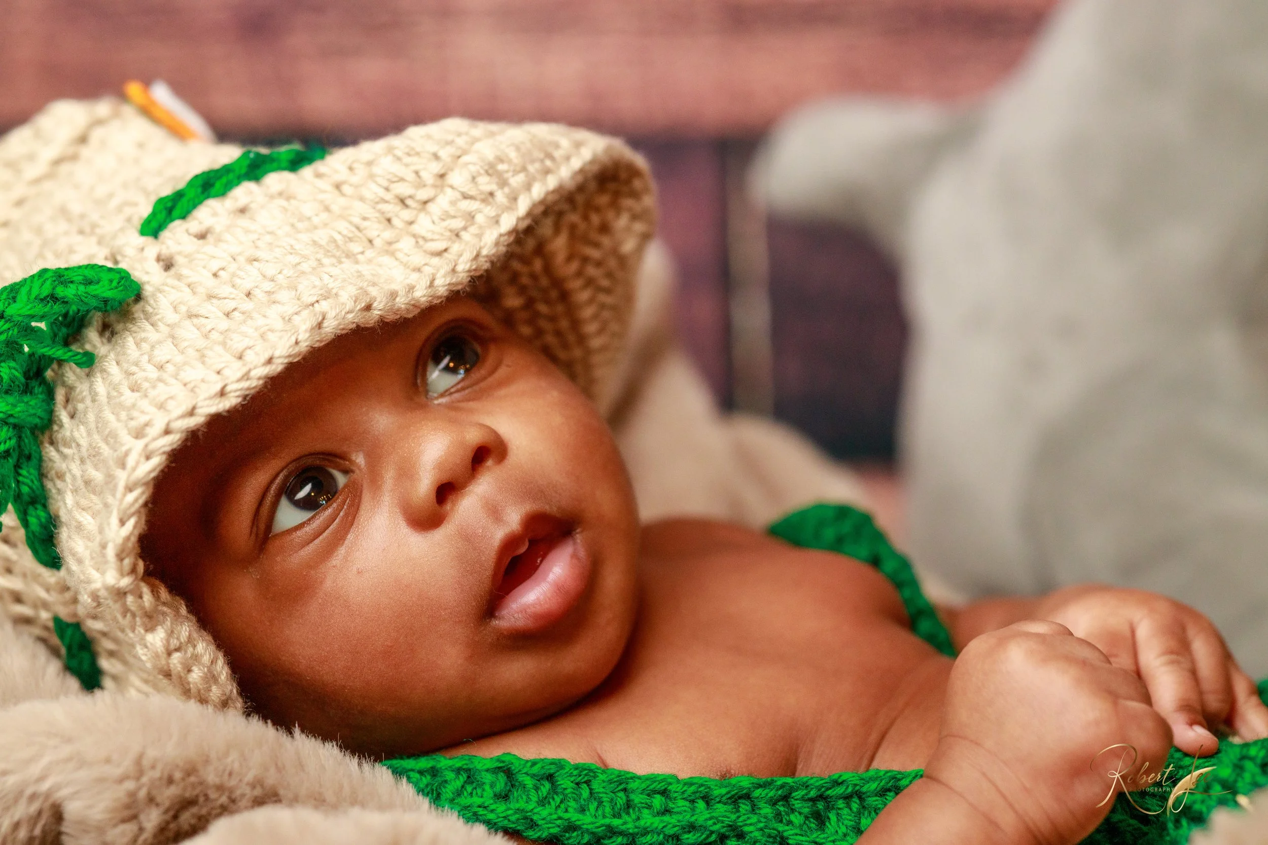 Close-up of a baby with a tan knitted hat with green details, lying on a soft blanket, gazing upwards.