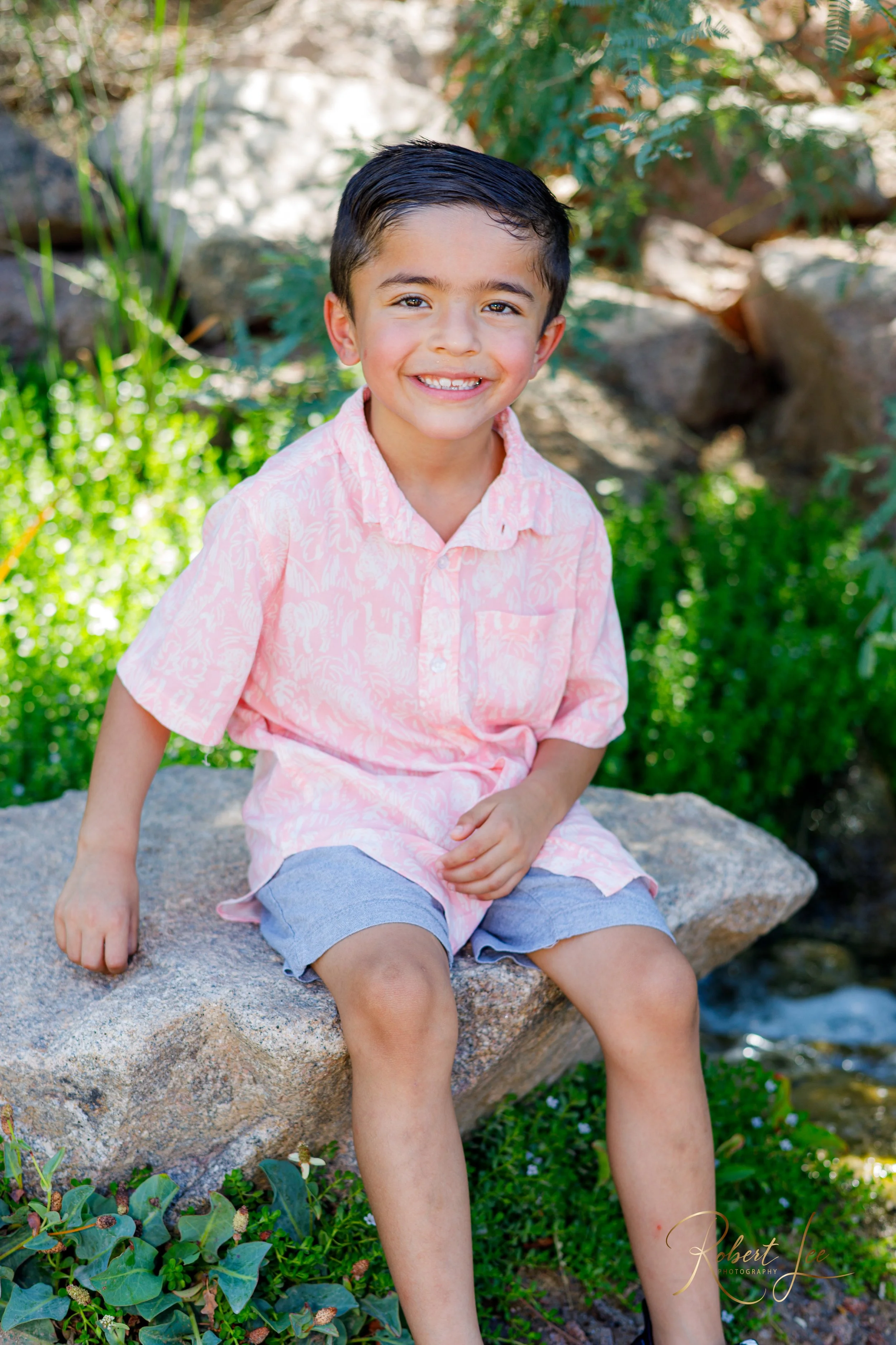 A young boy sitting on a large rock outdoors, smiling, wearing a light pink patterned short-sleeve shirt and gray shorts, with greenery and rocks behind him. Tucson Portrait photographer. Robert lee Photography.