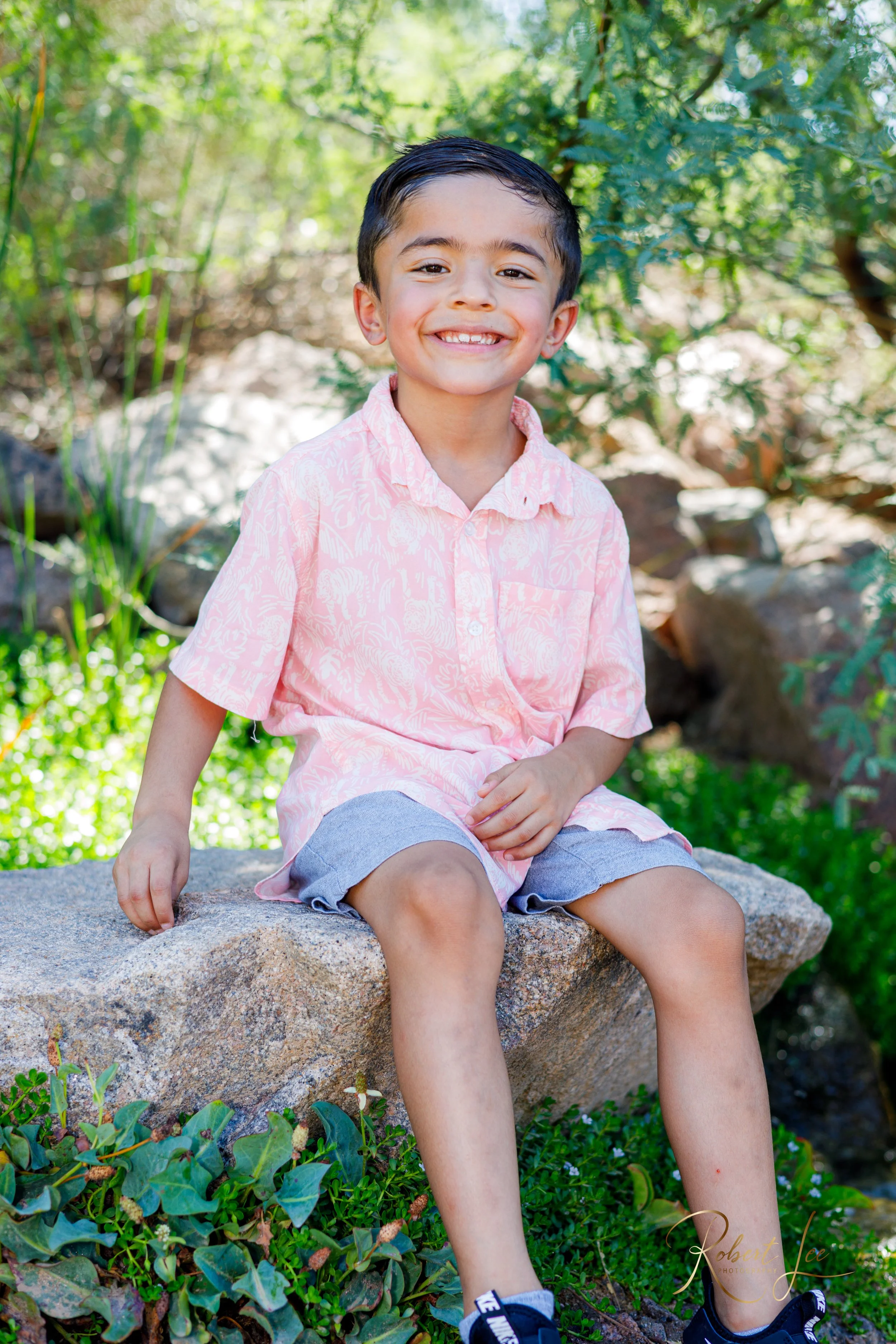 A young boy with dark hair is sitting on a large rock outdoors, smiling at the camera. He is wearing a pink patterned short-sleeve shirt, gray shorts, and black sneakers. Green plants and trees are in the background. Tucson Portrait photographer. Rob