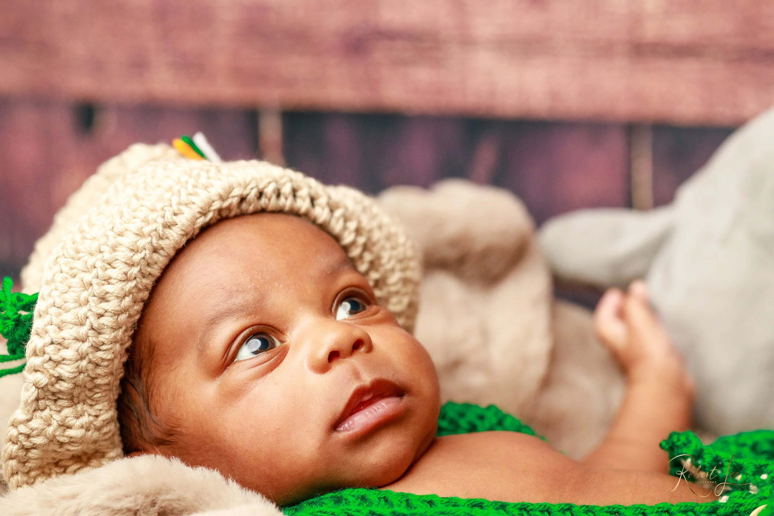 Close-up of a baby wearing a beige knitted hat with green accents, lying on a soft blanket, looking upwards.