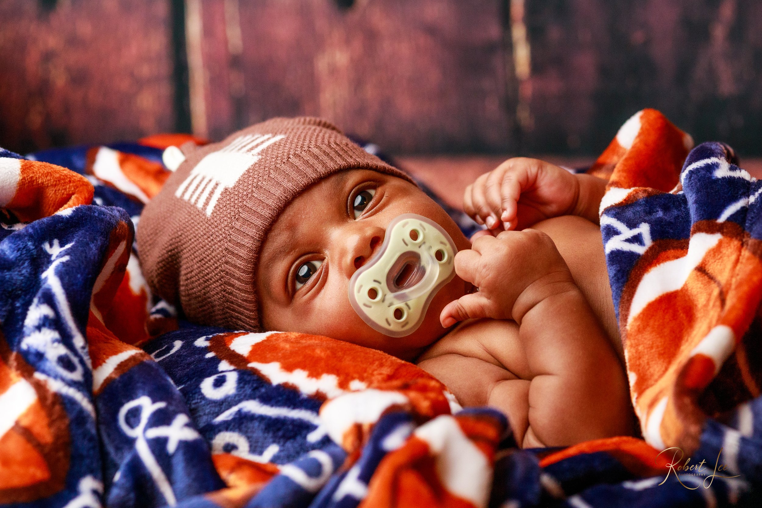 A baby lying on a colorful blanket, wearing a brown knit hat with a white design and a pacifier in their mouth.