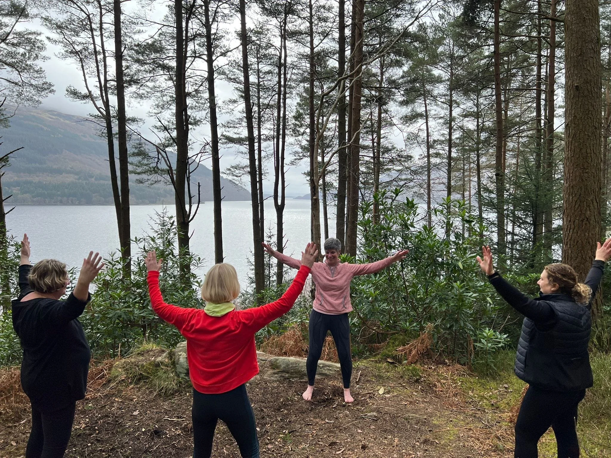 Four women in an outdoor forest setting near a lake, with one woman standing on the ground and others surrounding her, all raising their hands in a joyful pose.