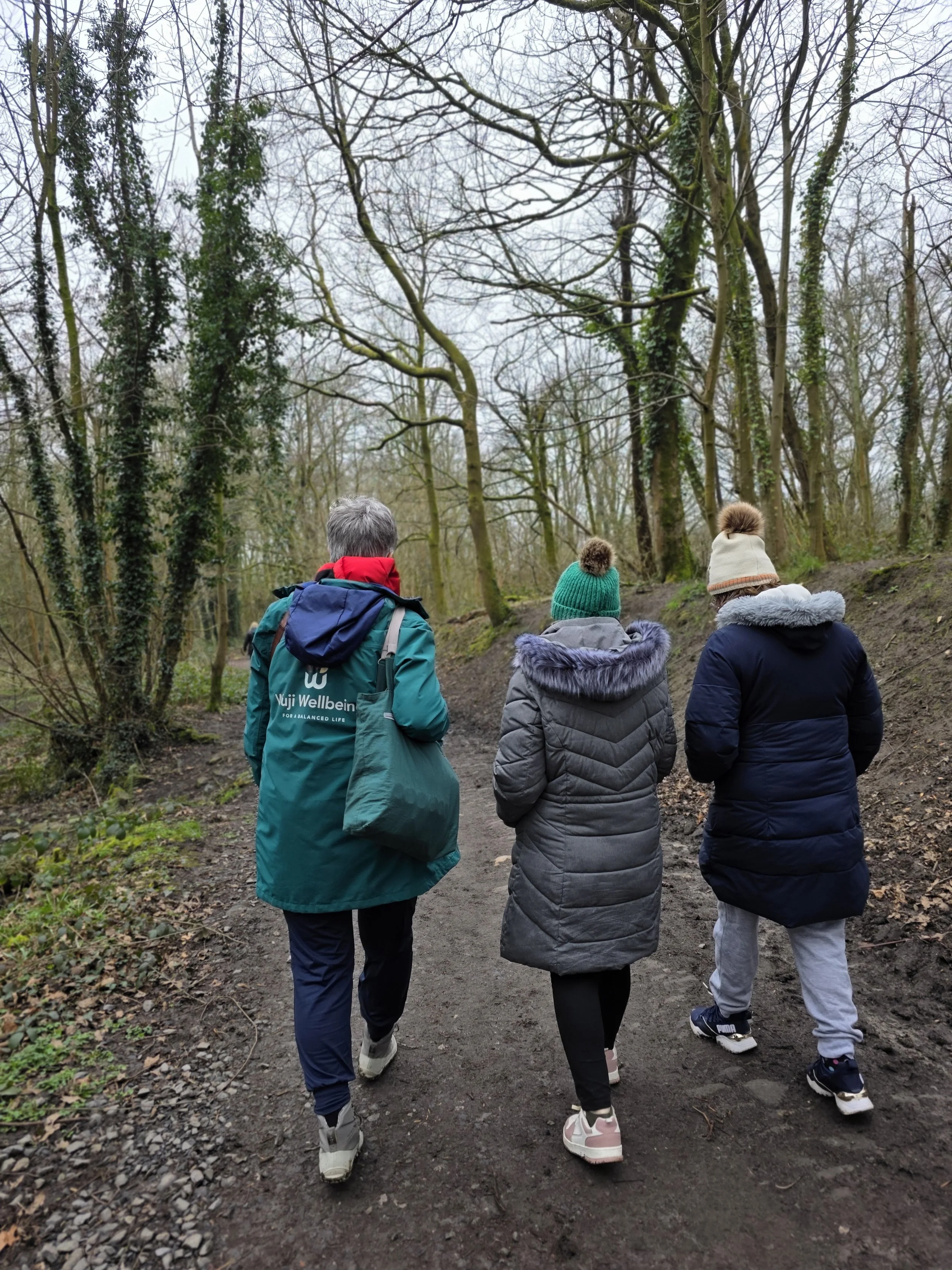 Qi Gong social side with Three people walking on a dirt trail in a forest, wearing winter jackets and hats.
