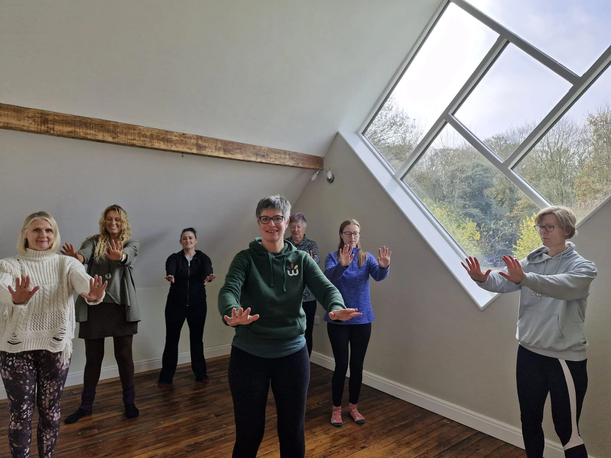 Seven women participating in a yoga or meditation session in a bright room with a large slanted window showing a view of trees outside. They are standing with their hands raised in front of them in a relaxed pose.
