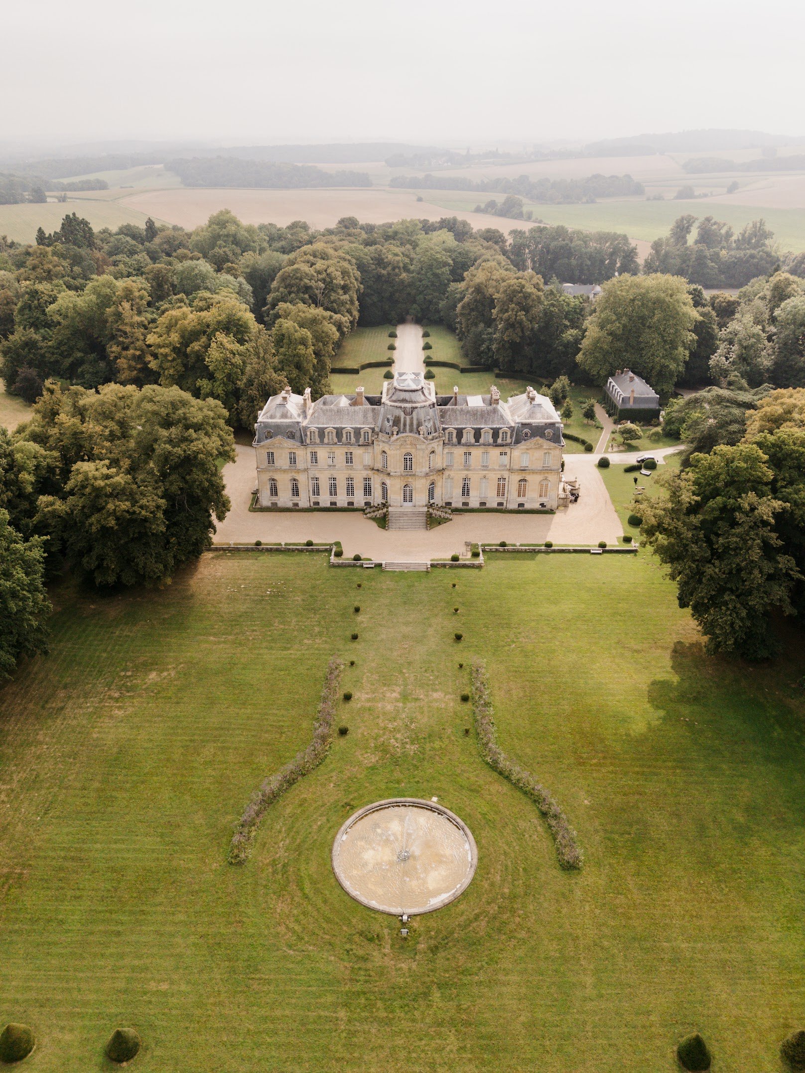 An aerial view of a large historic mansion - Chateau de Champlâtreux - surrounded by lush green trees and well-manicured lawns, with a circular fountain in the foreground and expansive farmland in the background.