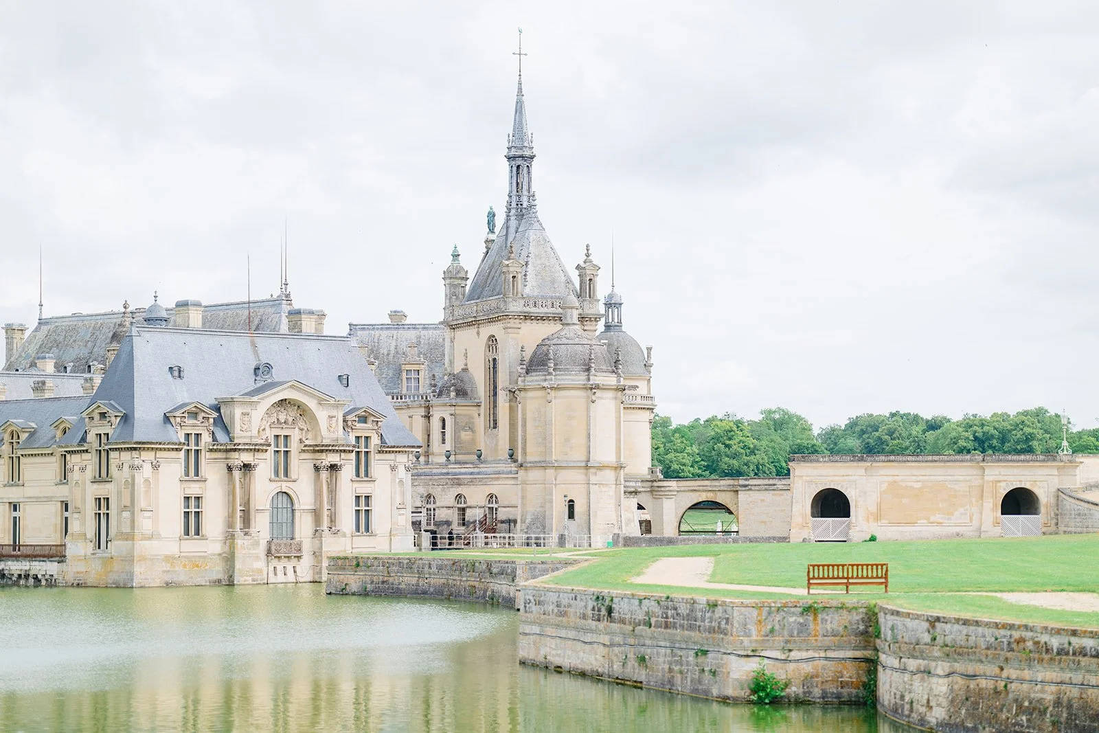 A historic castle with multiple towers and spires, situated by a moat with a wooden bench nearby, under a cloudy sky, surrounded by green trees in the background.