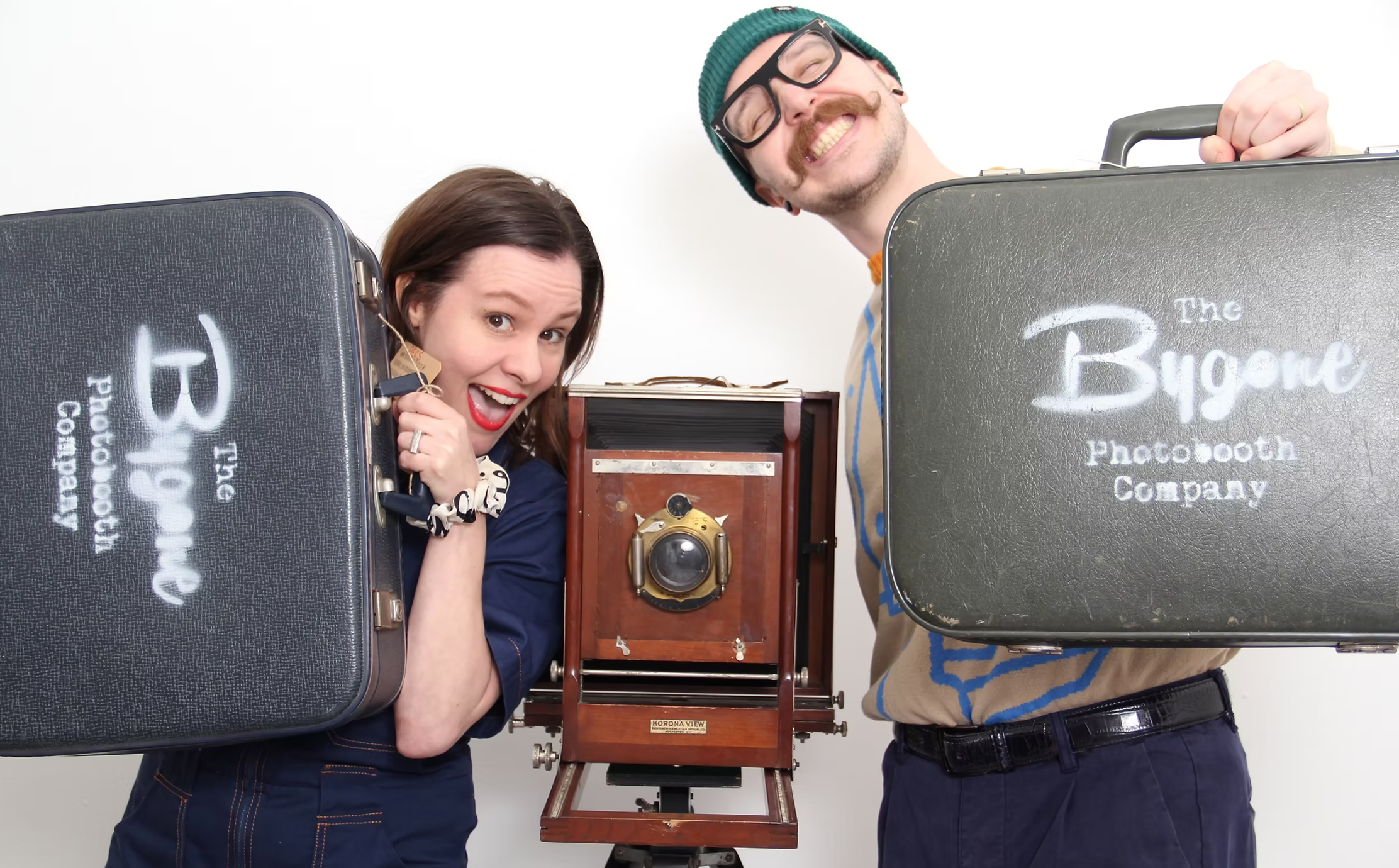 two people holding suitcases saying "the Bygone Photobooth Company" with a vintage camera between them