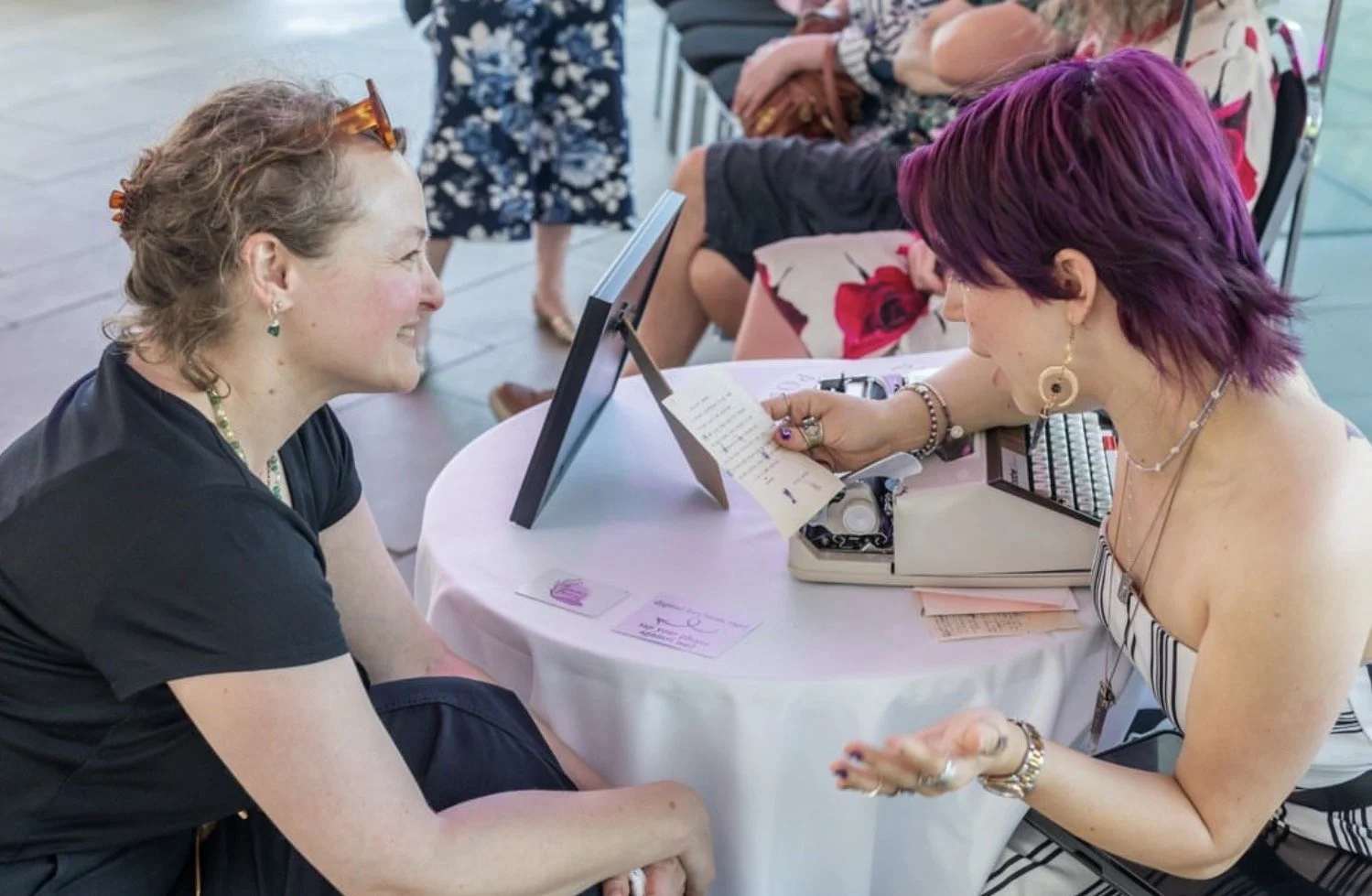 purple haired poet with typewriter reads poem to smiling guest