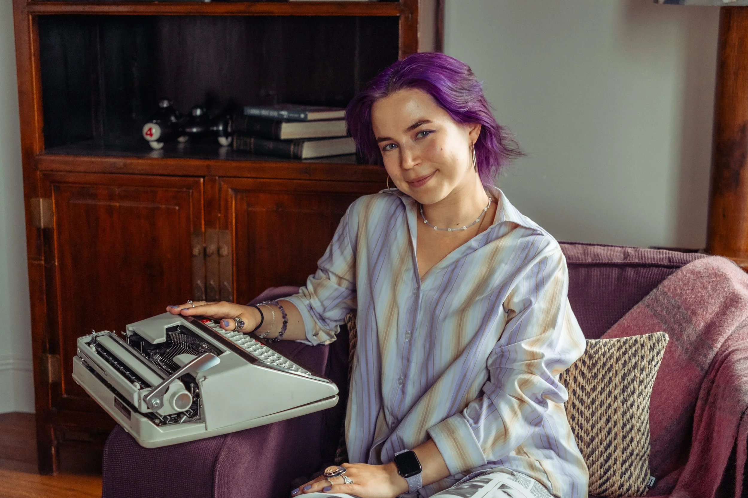 purple haired poet with typewriter sat in armchair smiling