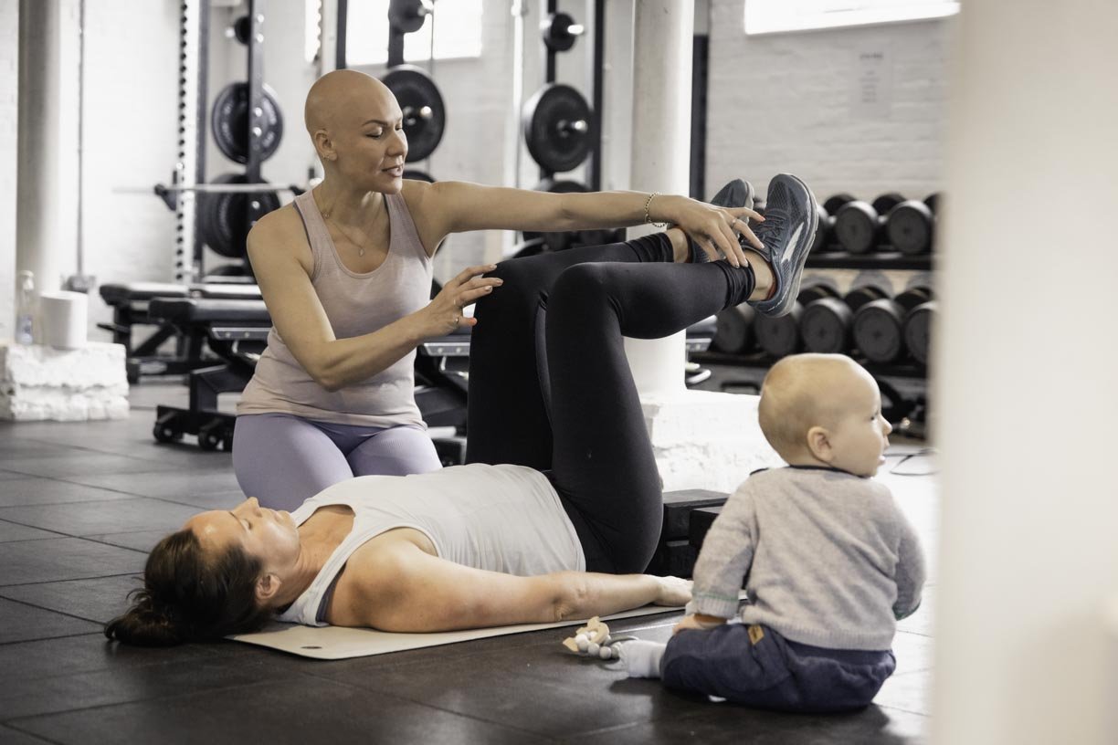 A woman is lying on a workout mat on the floor while another woman is helping her stretch her leg in a gym. The woman on the mat is wearing a gray tank top, and the woman helping her is dressed in a pink top and gray leggings. A young child with a bald head, wearing a gray sweater and dark pants, is sitting nearby, looking away. The gym has weights and equipment in the background.