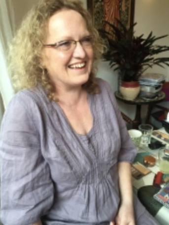 A smiling woman with curly blonde hair and glasses, sitting indoors with a cluttered table and a potted plant in the background.