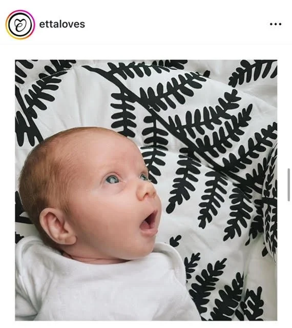 A surprised baby with blue eyes and light hair lying on a bed with black and white leaf-patterned bedding.