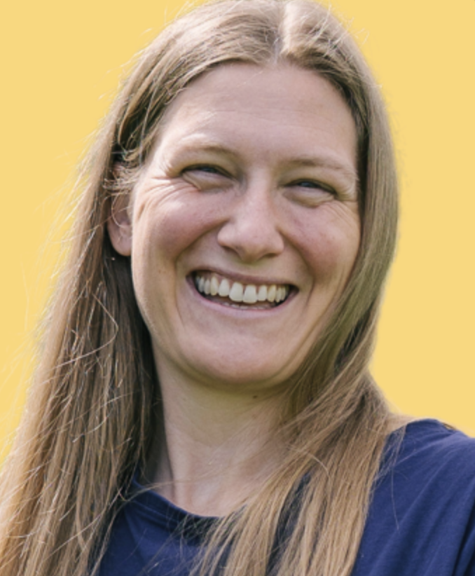 A smiling woman with long light brown hair, wearing a dark shirt, against a yellow background.